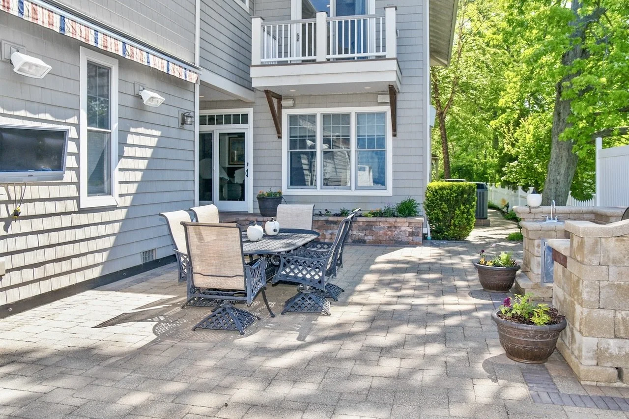 An outdoor patio area with a dining table and chairs, potted plants, a built-in outdoor sink, and a landscaped backyard with trees and bushes.