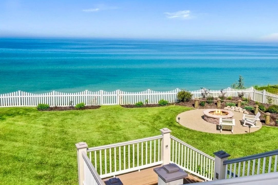 View of a landscaped backyard with green grass, garden beds, white picket fences, and a round patio with chairs, overlooking a calm blue ocean under a clear sky.