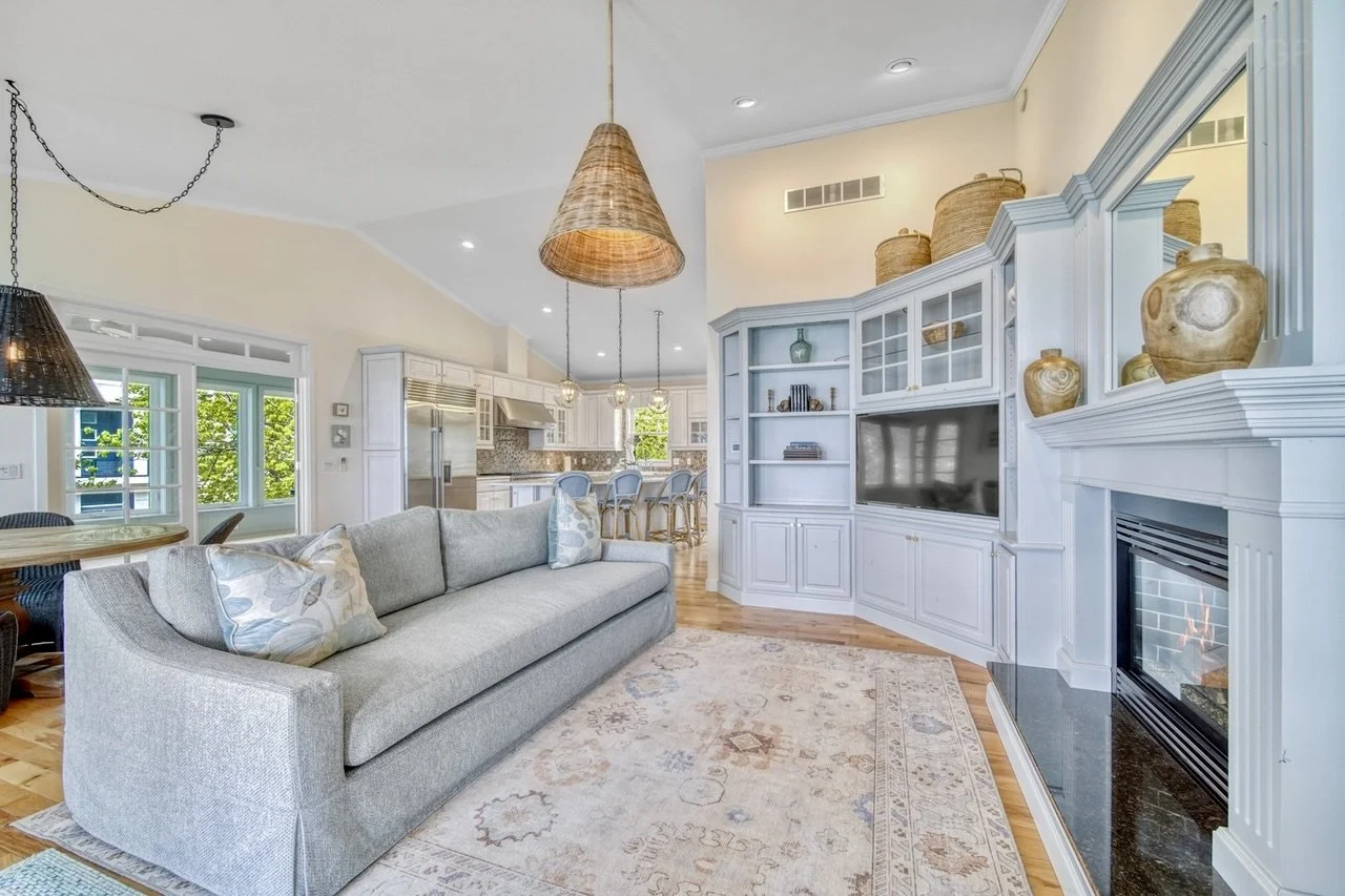 Living room with a grey sofa, patterned pillows, a white built-in entertainment center with a television, a fireplace with a black hearth, and decorative vases on the mantle, adjacent to a dining area and open kitchen with bar stools and large windows bringing in natural light.