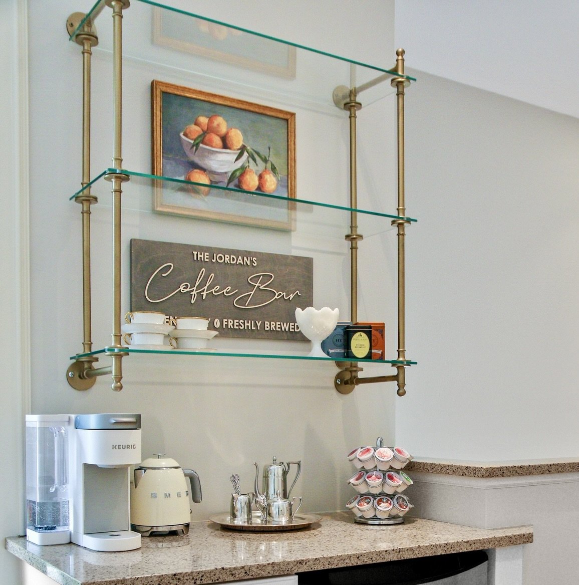 Kitchen coffee station with Keurig machine, tea kettle, cups, and coffee pods on a counter, with wall art and a sign that reads 'The Jordan's Coffee Bar'.