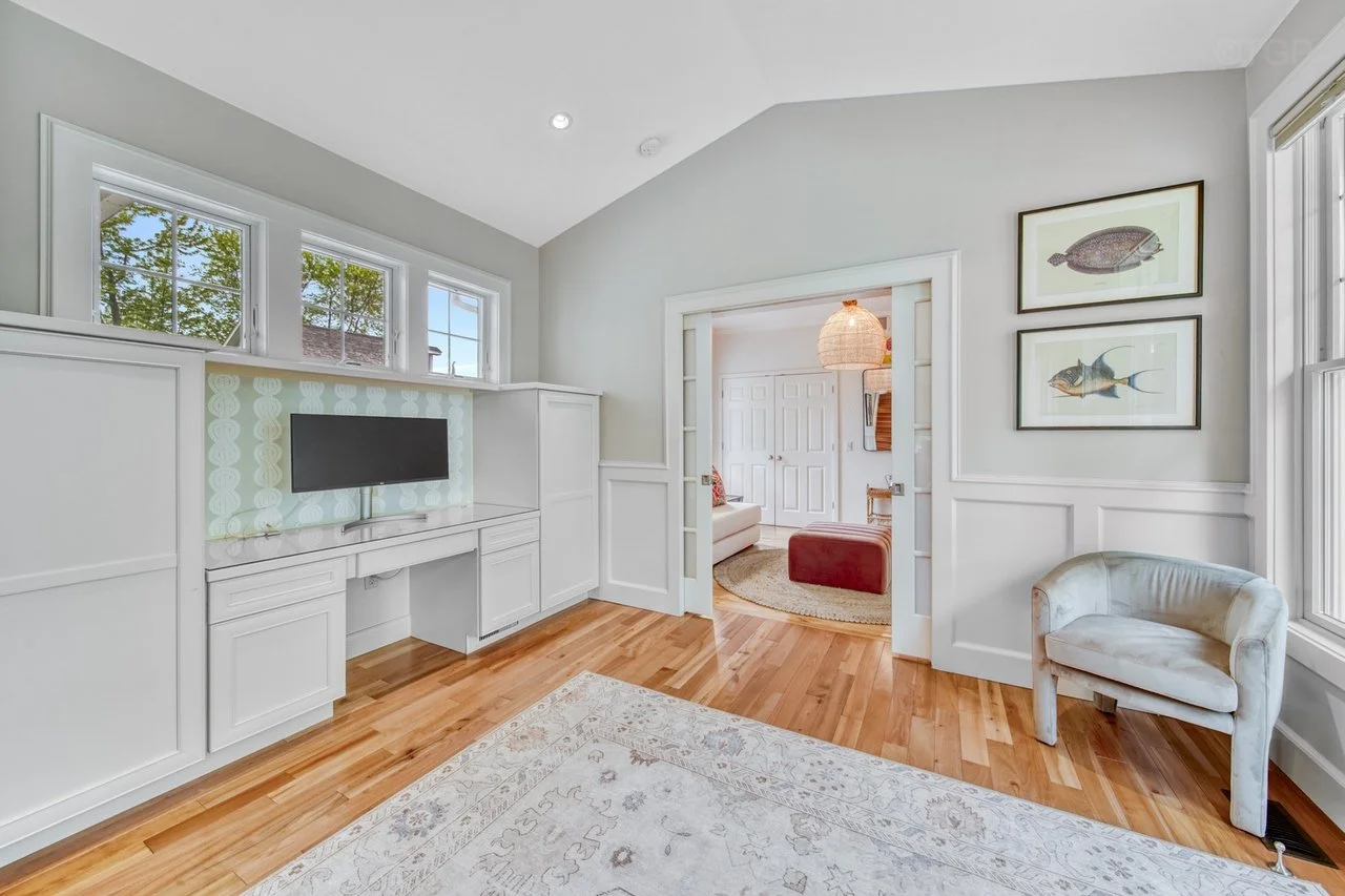 Living room with white built-in cabinets, a flat-screen TV, three windows, artwork of fish on the wall, hardwood floor, and a white and gold chair.