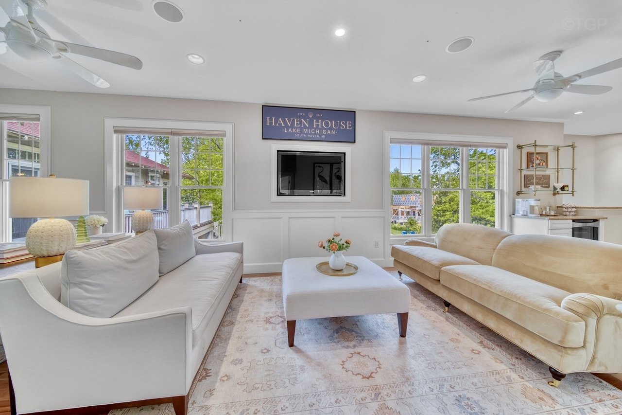 Bright living room with white and beige sofas, a coffee table with a flower vase, large windows showing greenery outside, wall-mounted TV, and a sign that reads 'Haven House Lake Michigan.'