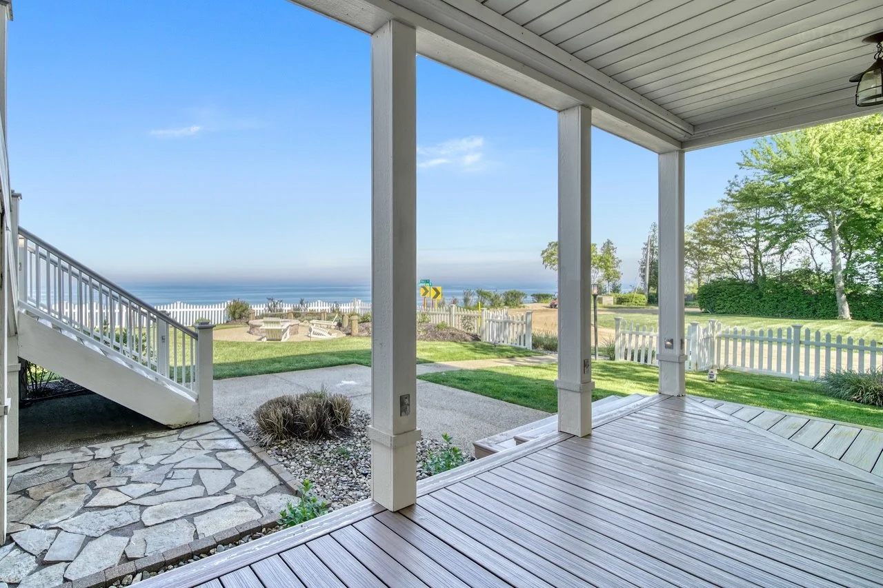 View from a covered porch looking out to a lawn with a white fence, trees, and Lake Michigan, under a clear blue sky.