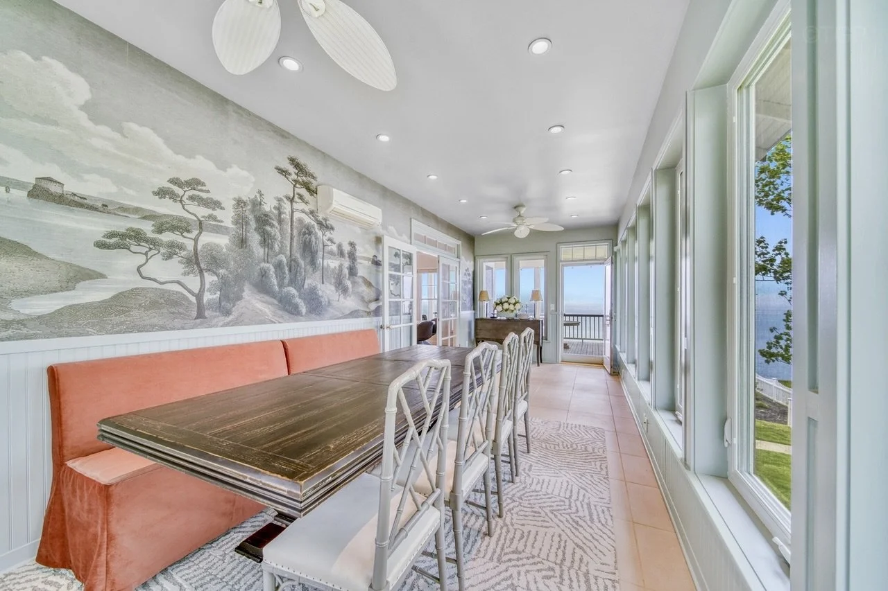 Sunroom with large windows, wooden dining table, coral bench, and white chairs. Wall mural of a landscape with trees and water. Ceiling fans and lamps, with an outdoor view of a deck and lake.