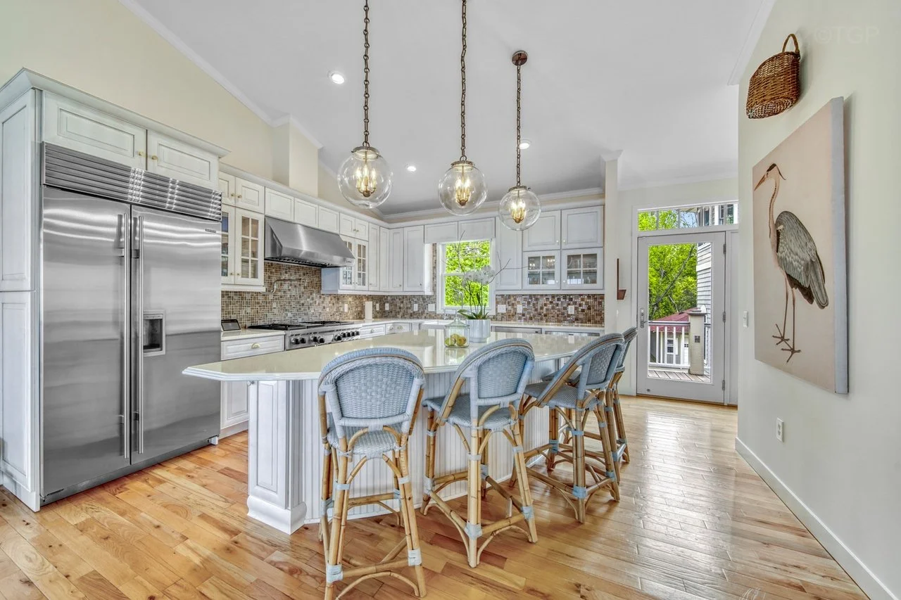 A bright kitchen with white cabinets, a large stainless steel refrigerator, a stovetop with a range hood, mosaic tile backsplash, a white island with a curved edge, and four wicker bar stools with blue cushions. There are three glass pendant lights hanging above the island and a window and glass door leading outside. Decorative elements include a painting of a bird, a basket, and a potted plant on the island.