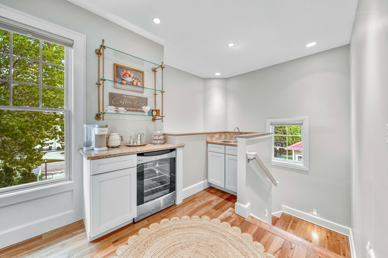 A small kitchenette with white cabinets, a mini wine cooler, granite countertops, and a window showing green trees outside.