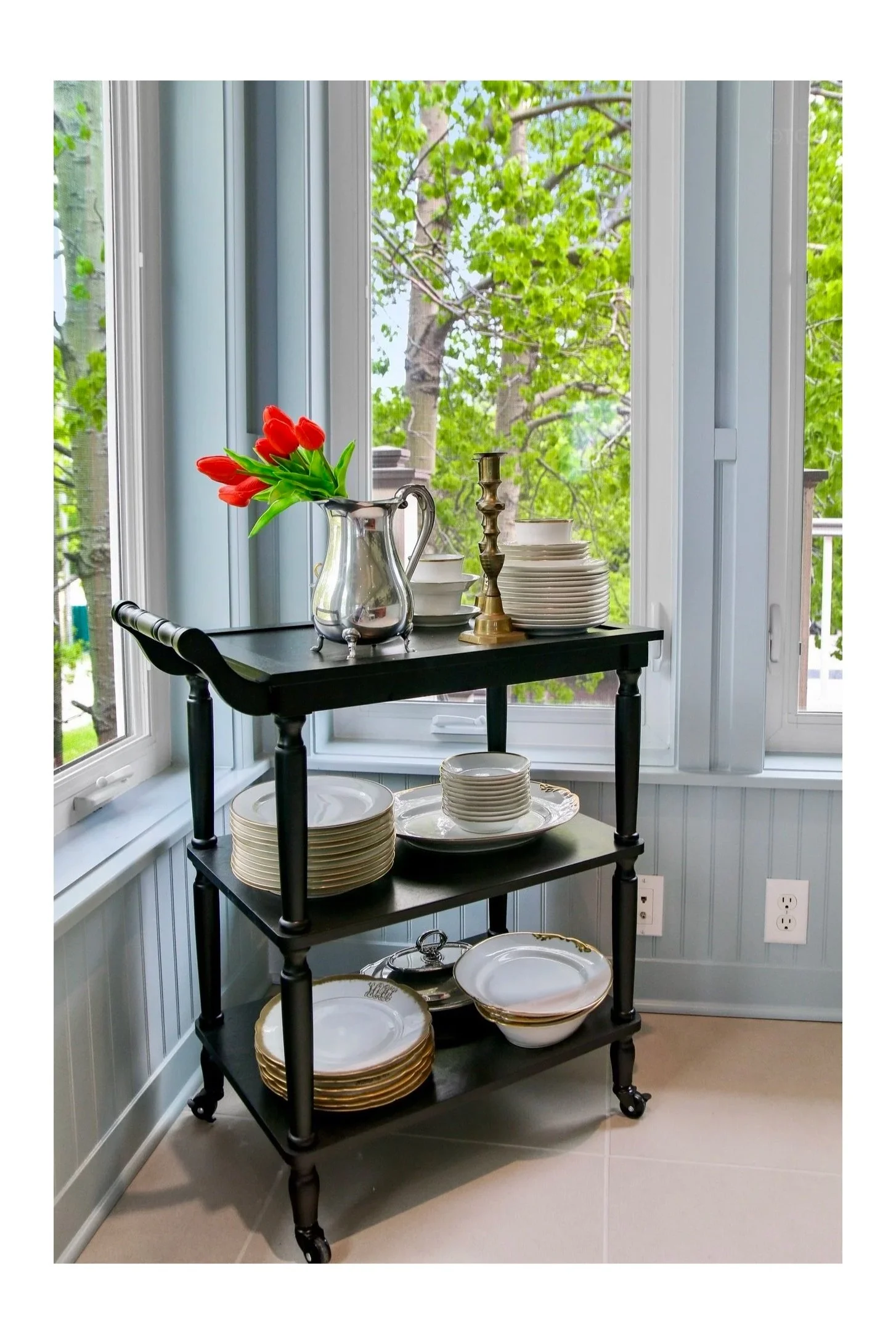 A black tiered serving cart with plates, bowls, a glass pitcher with red tulips, brass candlesticks, set in front of a large window with green trees outside.