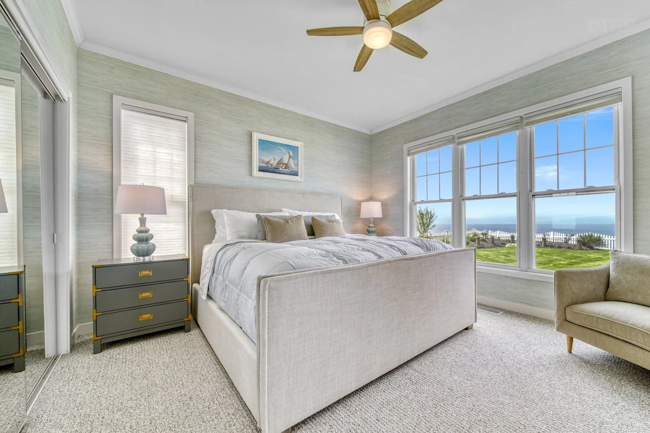 Bright bedroom with large windows showing ocean view, white bed with beige headboard, side tables with lamps, beige armchair, ceiling fan, and closet with mirrored sliding doors.