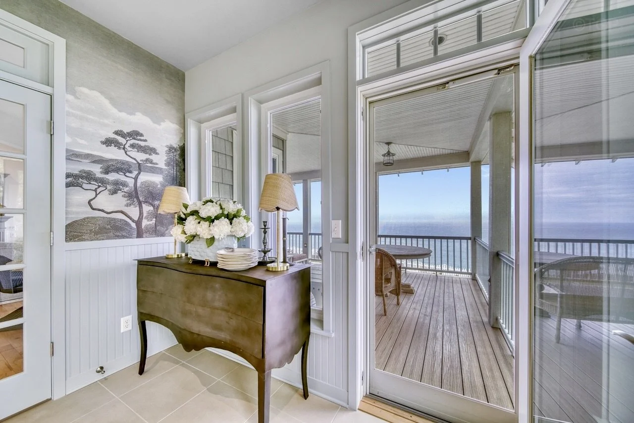 Interior view of a room with a wooden console table holding a vase of white flowers, two lamps, and a stack of plates. The room opens to a screened porch with a round table and chairs, overlooking the lake.