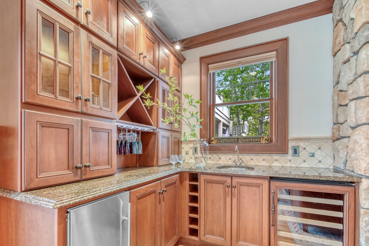 A cozy home bar area with wooden cabinets, granite countertops, a wine fridge, and a window with a view of trees outside. A small sign on the windowsill reads 'Captain's Quarters'.