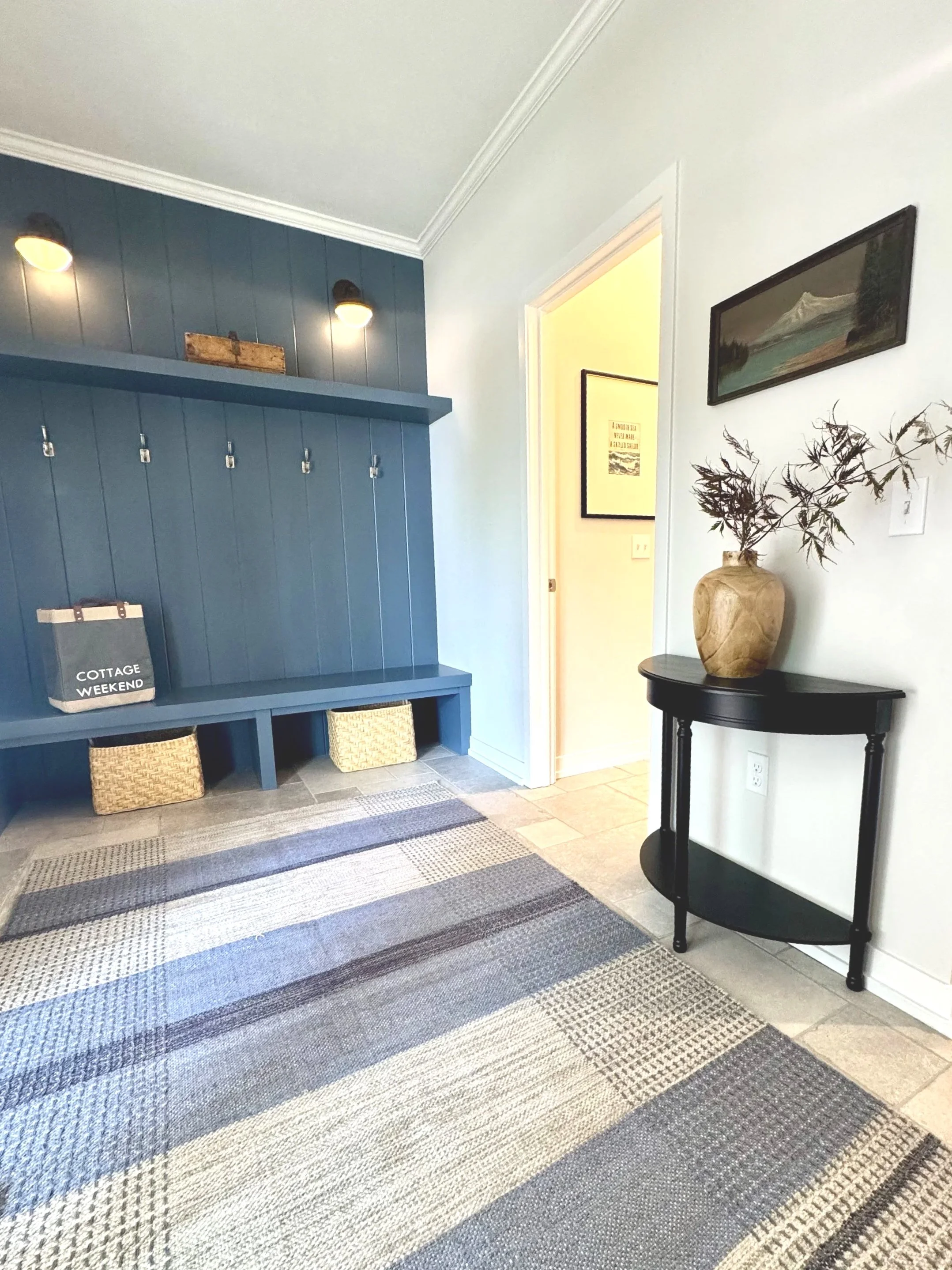 Entryway corner with a blue wooden hall tree, wicker baskets underneath, a wall art piece, a black side table with a large clay vase and decorative branches, and a striped area rug on tiled floor.