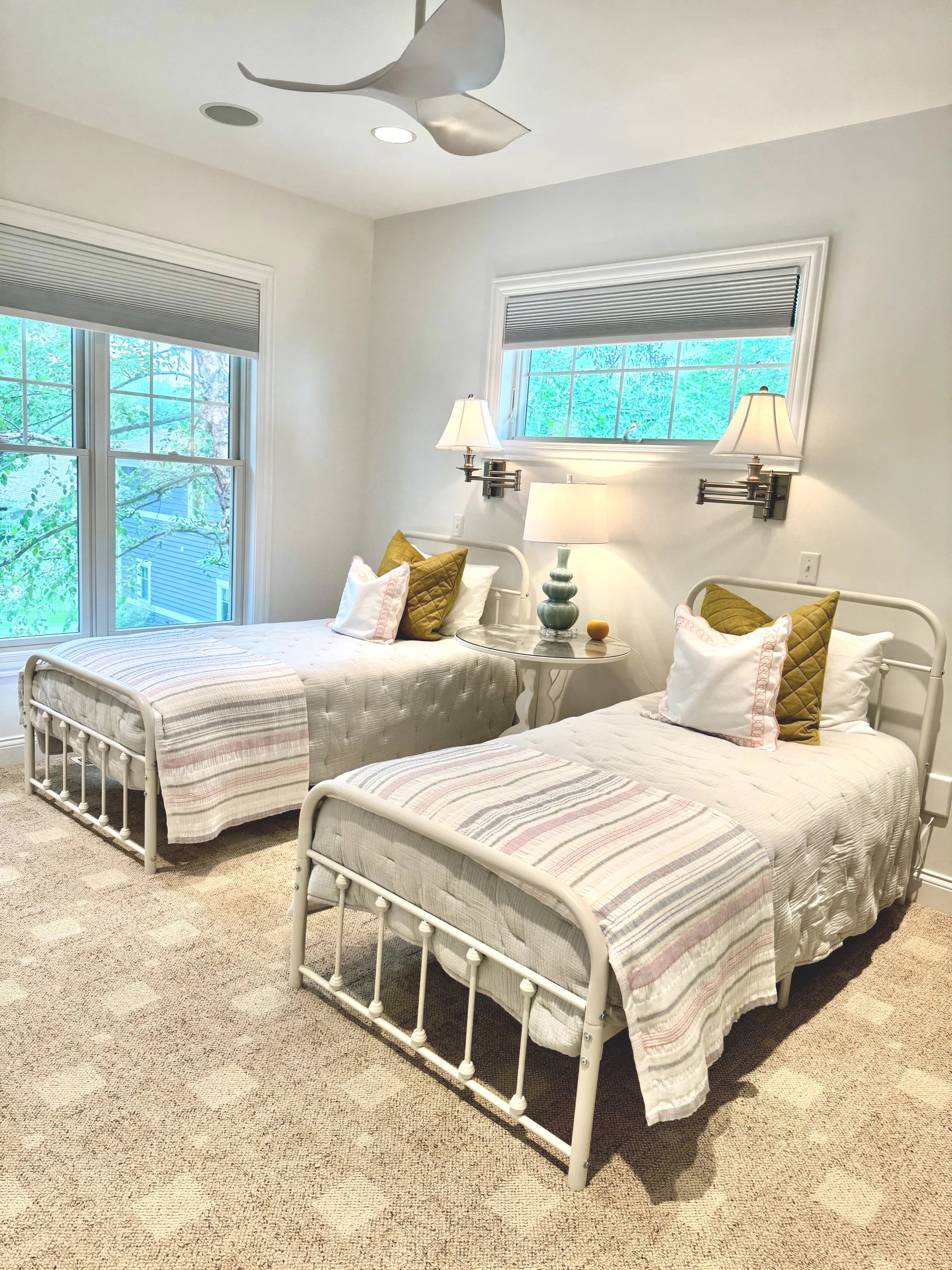 A bedroom with two twin beds, each with white metal frames and striped quilts. There are pillows and mustard-colored accent pillows on each bed. Between the beds is a round bedside table with a lamp. Above each bed is a wall-mounted lamp, and a horizontal window with blinds is above the beds. The room has large windows showing trees outside, and beige carpeted flooring.