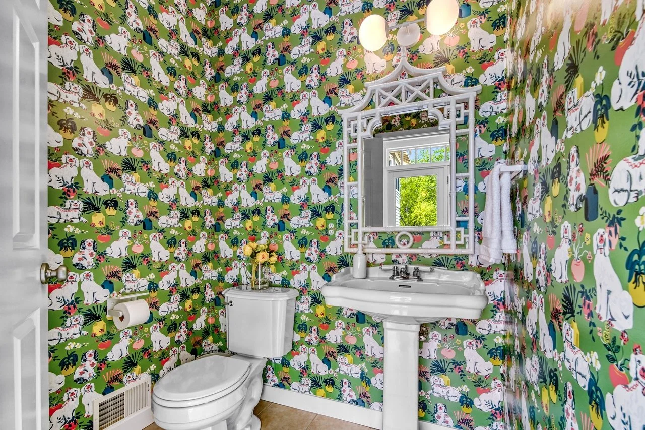 Bathroom with colorful wallpaper featuring white dogs, plants, and playful patterns, white porcelain toilet, pedestal sink with a mirror above, small window showing greenery outside, towel hanging on the wall, and a vase with flowers on the toilet tank.