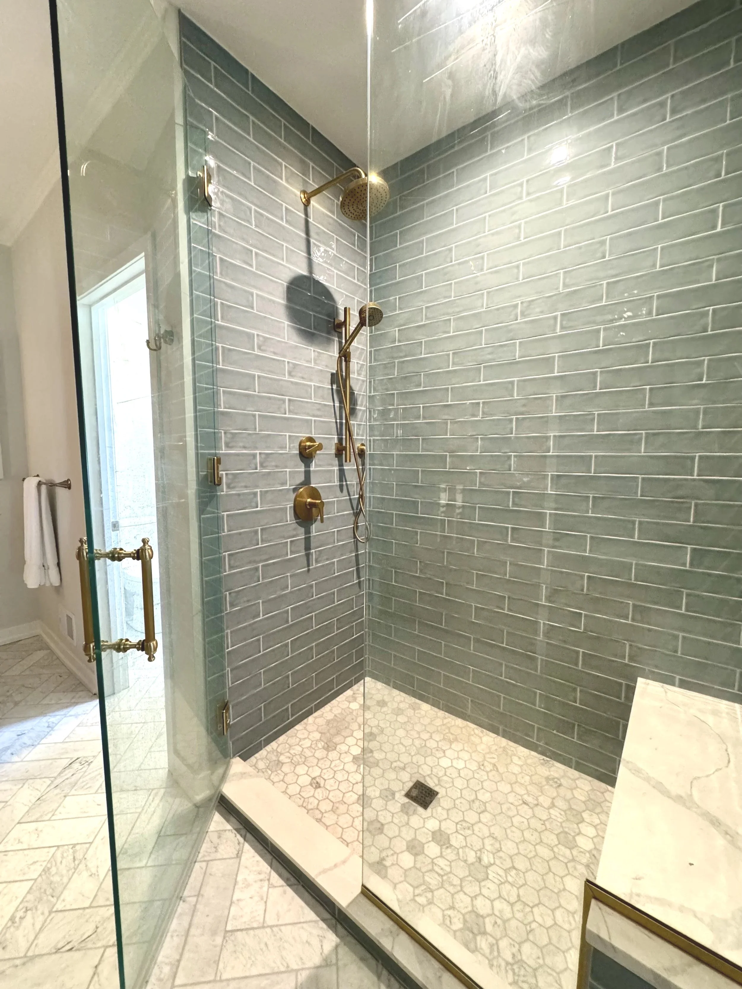 A modern walk-in shower with dark gray subway tile walls, brass fixtures, and a glass enclosure with gold hardware. Inside the shower, there are two showerheads and a built-in bench. The shower floor has small hexagon tiles and a drain in the center.