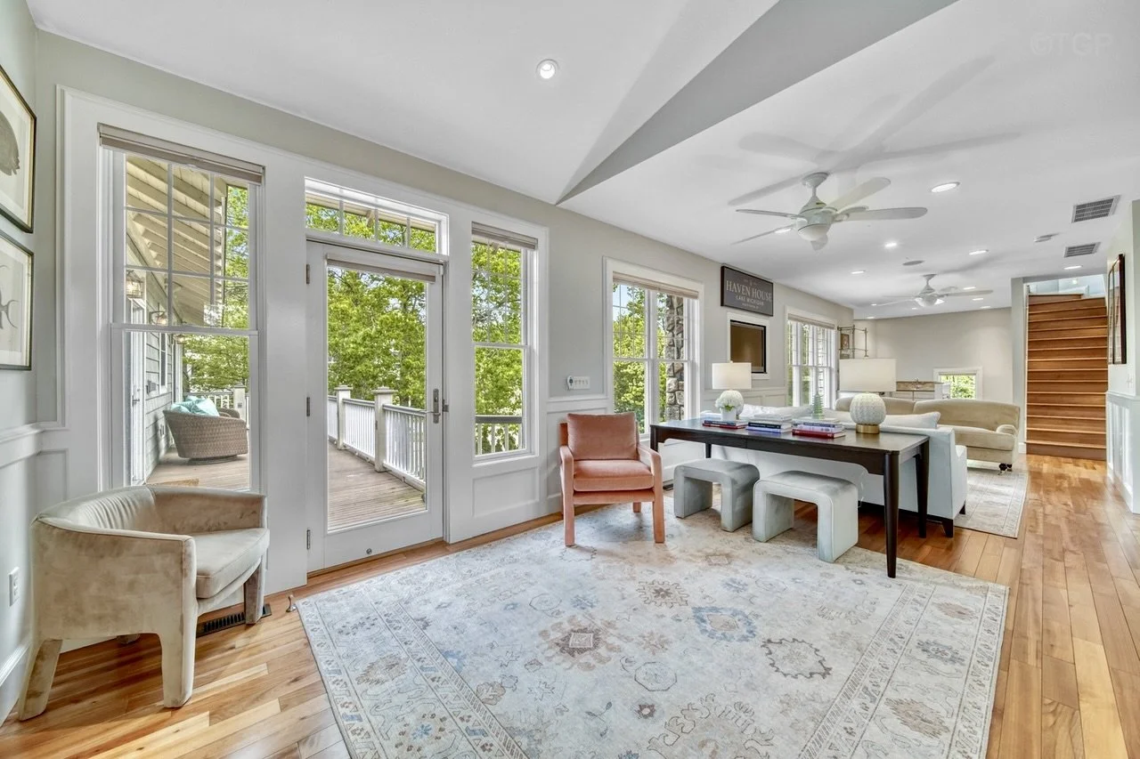 Bright living room with large windows, wooden floors, white walls, and a view of a deck outside. There are various chairs and couches, a white rug, a dark dining table with stone stools, and some lamps.