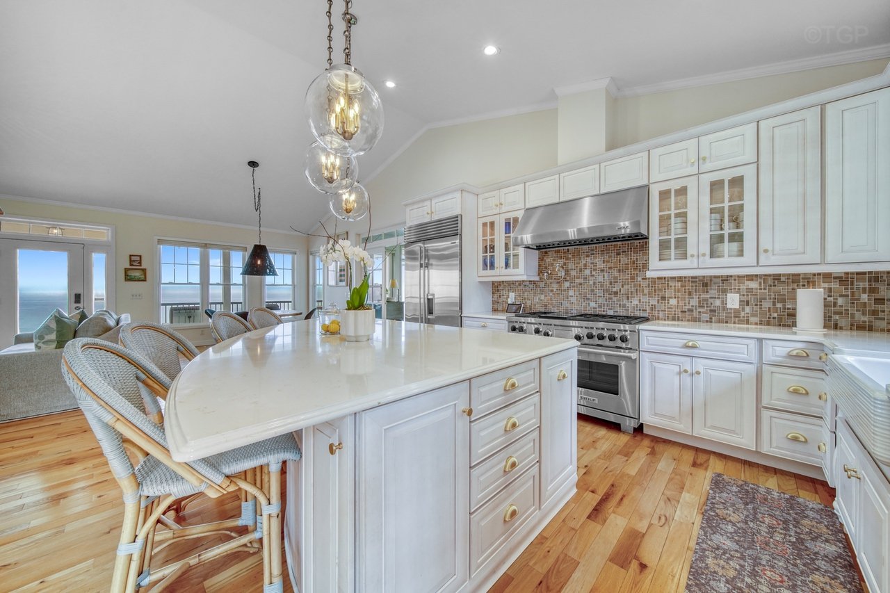 Bright kitchen with white cabinets, a large island with seating, a stainless steel stove and refrigerator, wooden floors, and large windows showing a view outside.
