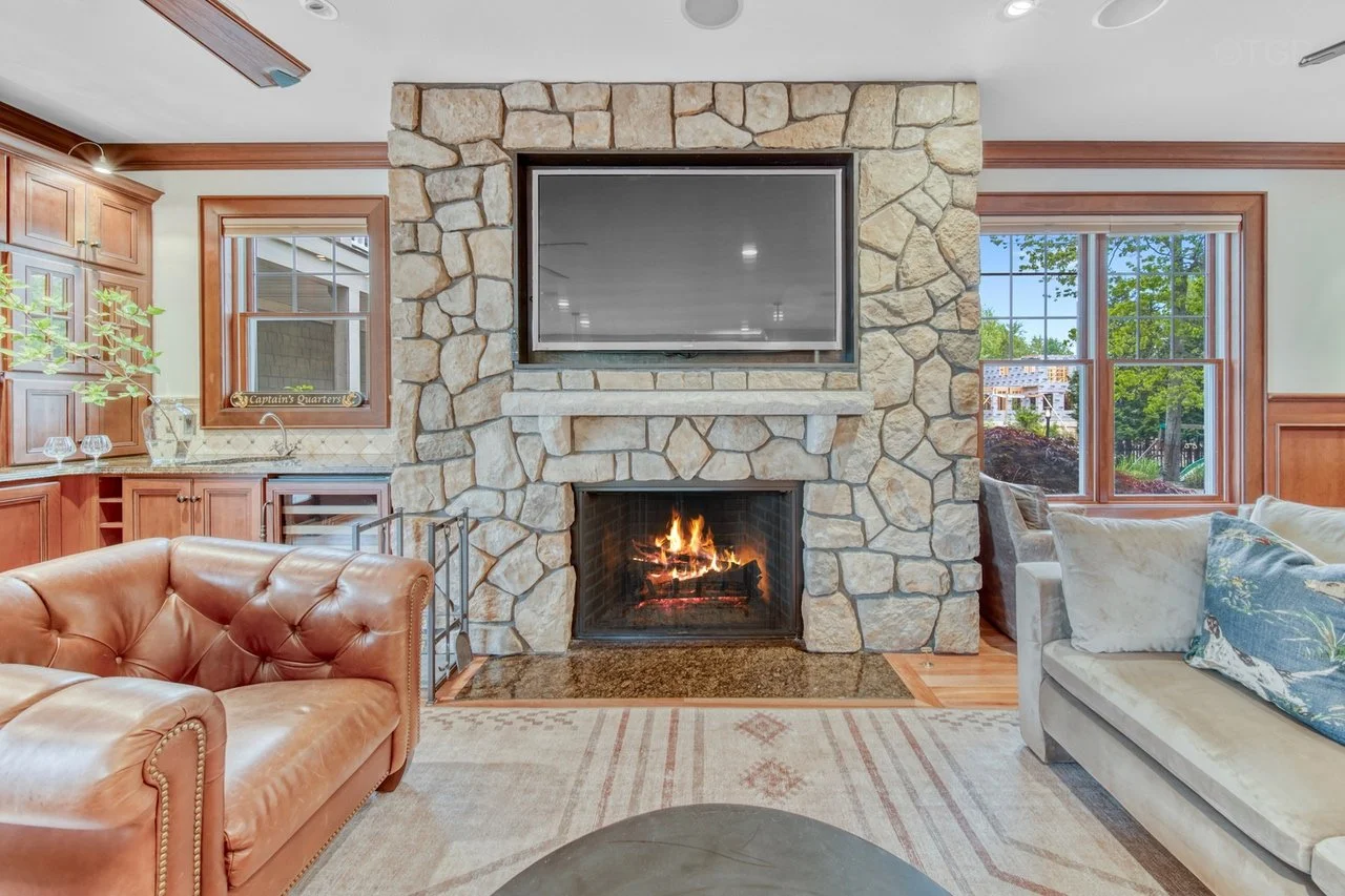 Living room with stone fireplace, mounted television, leather armchair, and sofa near windows with trees outside.