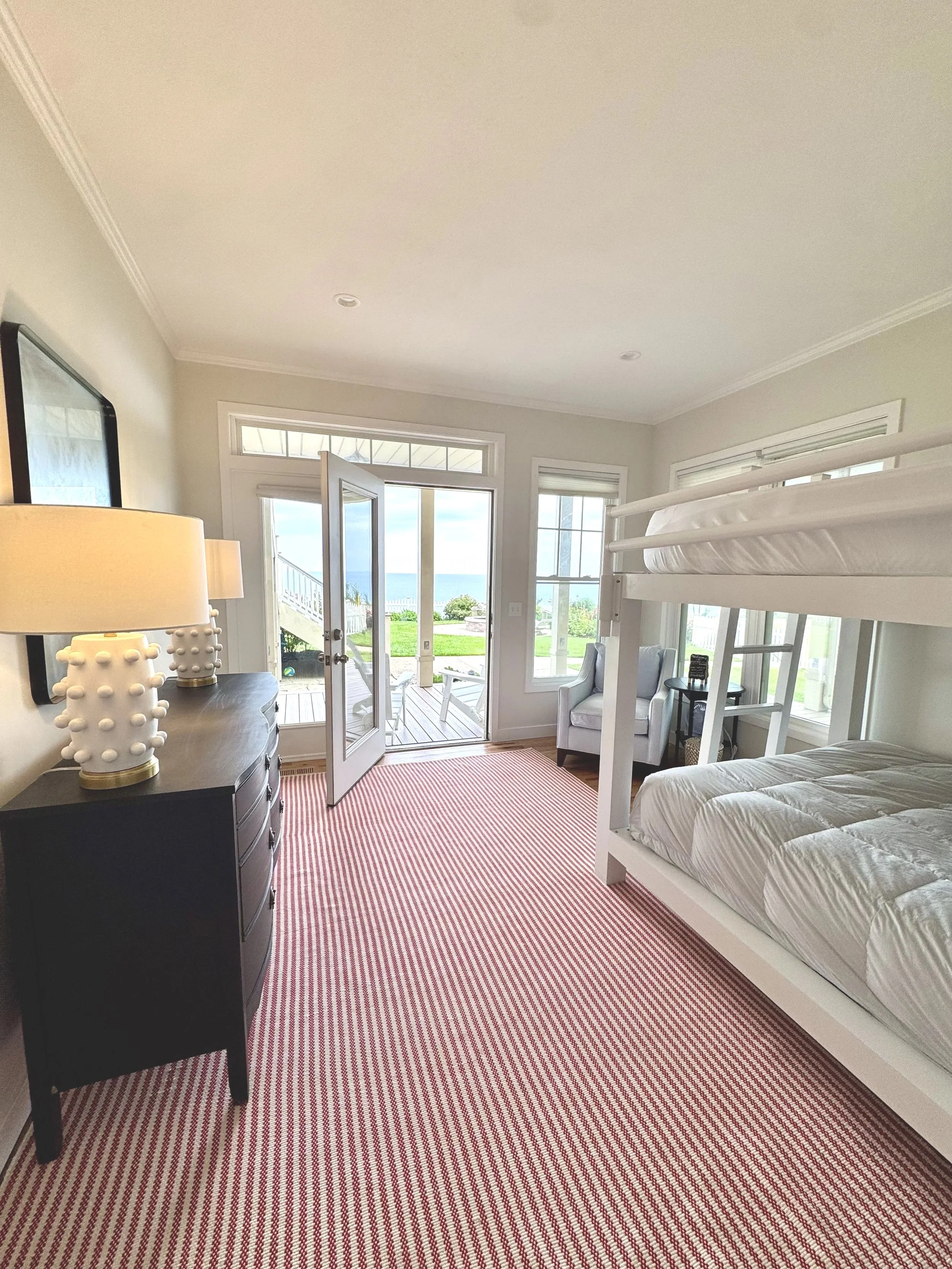 A bedroom with large windows and a view of the ocean. It has a white bunk bed, an armchair, a black dresser with decorative lamps, and a sliding glass door leading to an outdoor porch.
