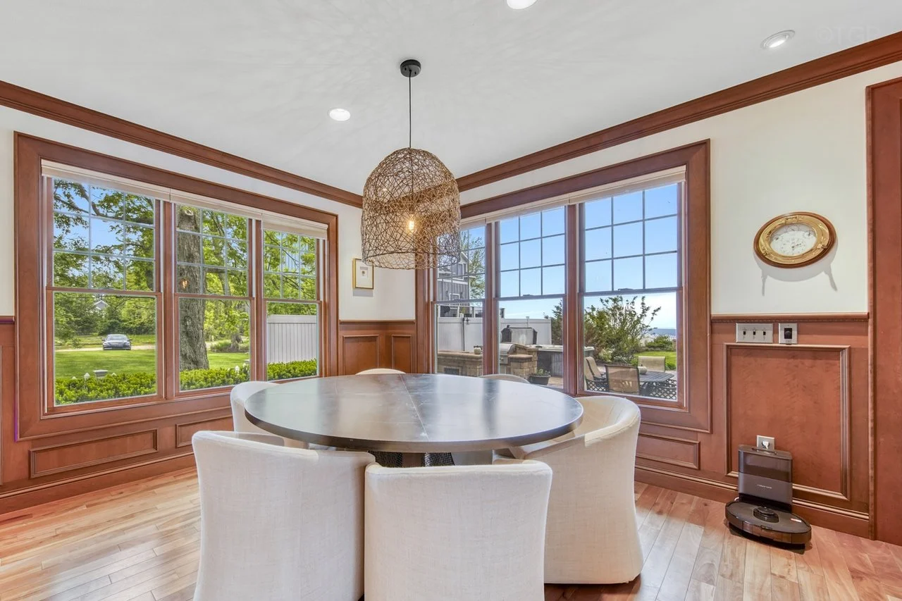 Dining room with large windows, wooden trim, round table, and cushioned chairs, overlooking a yard with green grass and trees.