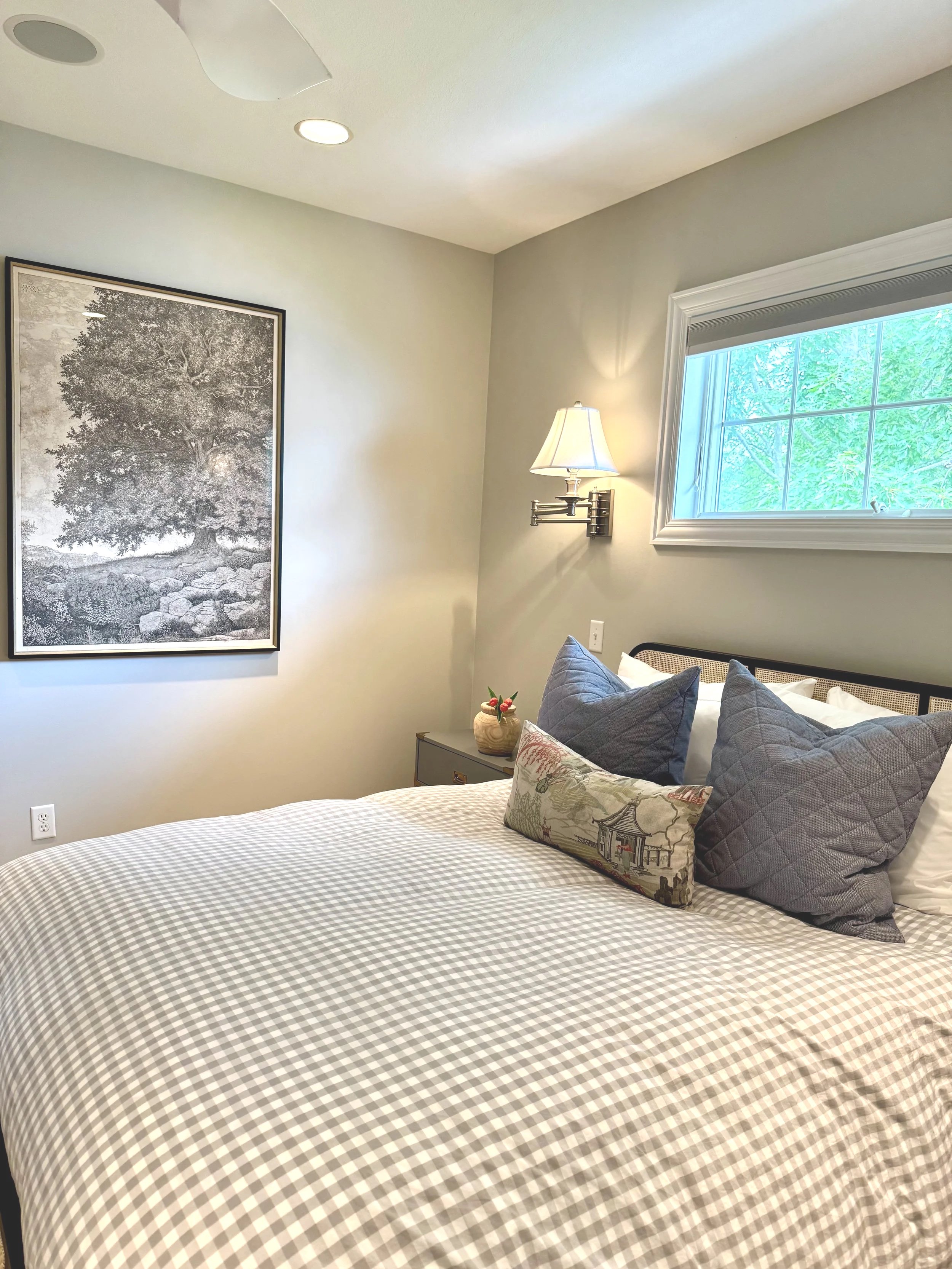 A neatly made bed with a checkered bedspread, dark gray pillows, and a decorative lumbar pillow with a landscape and architectural scene, in a bedroom with a window, wall art, and wall-mounted lamp.