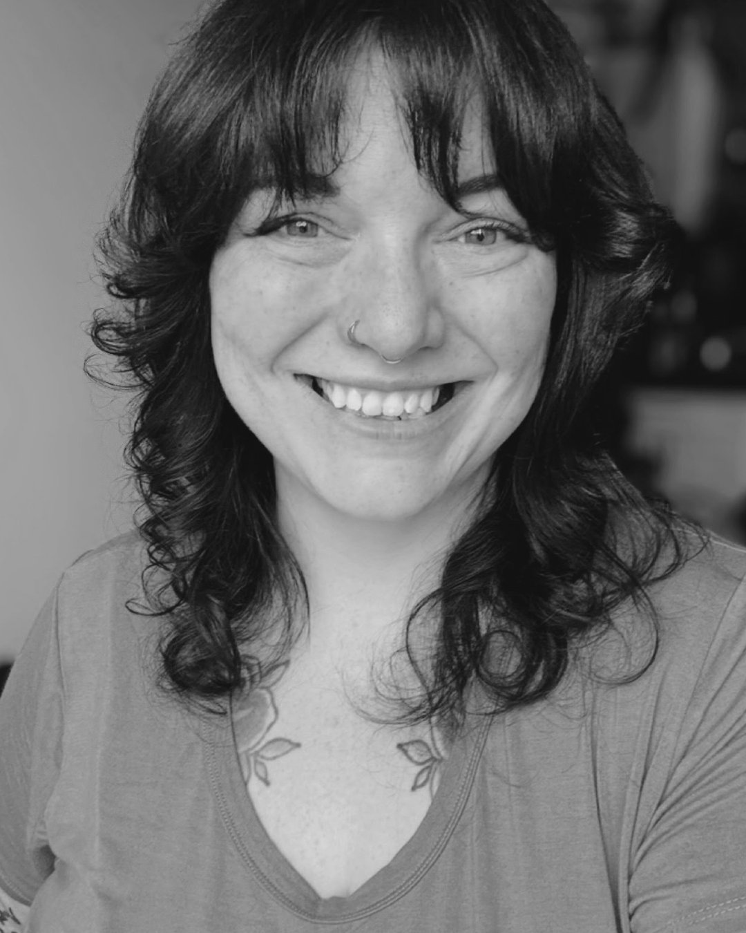 Black and white close-up photo of a smiling woman with wavy dark hair, light-colored eyes, a nose piercing, and tattooed shoulders.