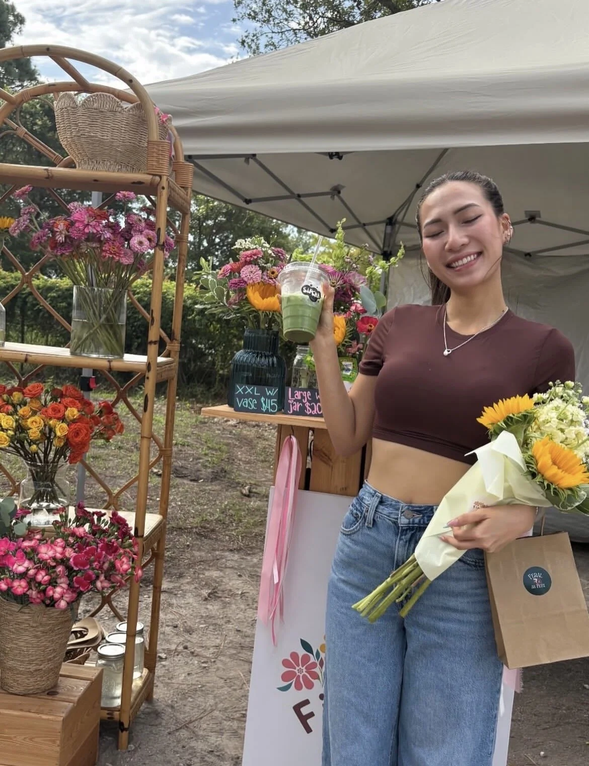 A woman at a flower stand holding a bouquet of sunflowers and white flowers, smiling with closed eyes, wearing a maroon crop top and blue jeans, holding a green smoothie, with various colorful flower bouquets on display under a canopy.