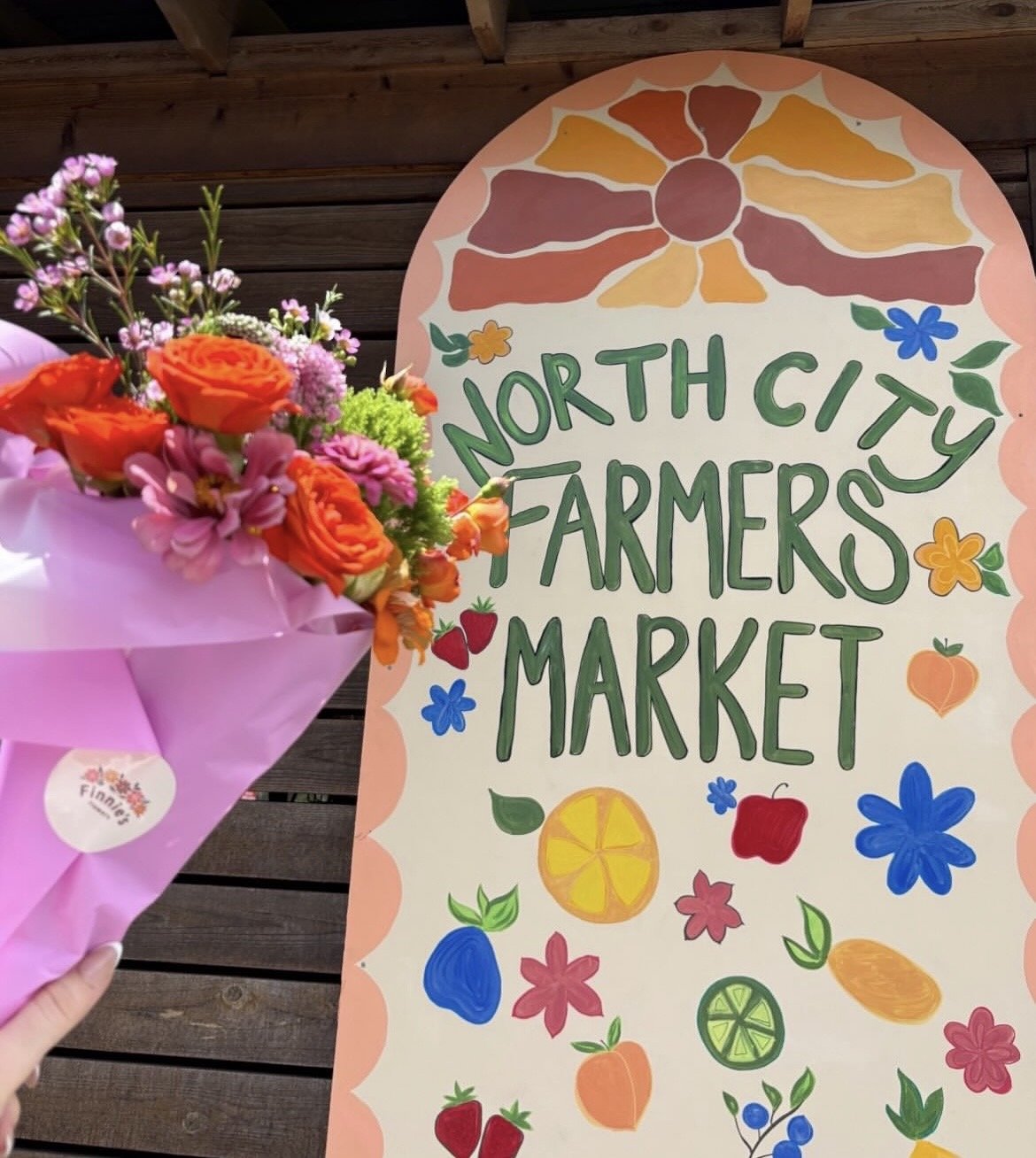 A bouquet of brightly colored flowers held by a person with a pink wrapping paper, placed in front of a sign for North City Farmers Market decorated with colorful fruits and flowers at a wooden backdrop.