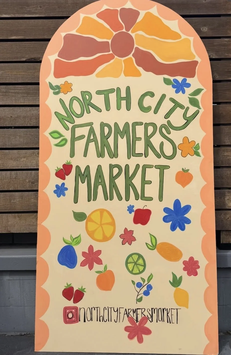 A colorful sign for North City Farmers Market with hand-drawn fruits, vegetables, and flowers, featuring a sunset illustration at the top.