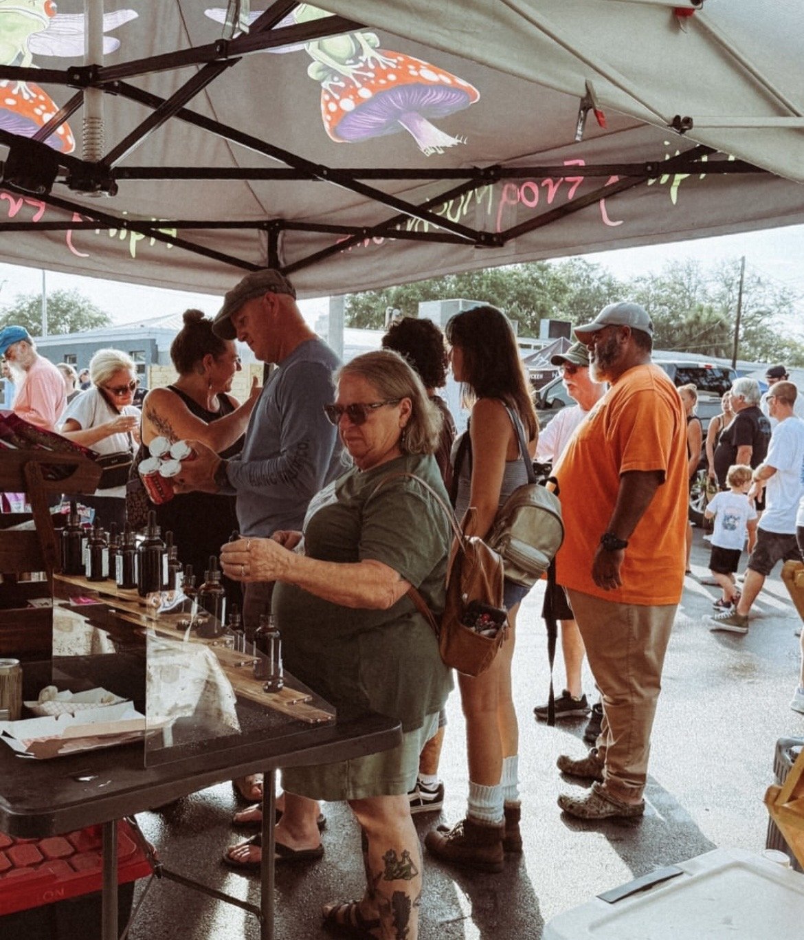 People standing in line at an outdoor market tent, browsing and shopping for small bottles and products.