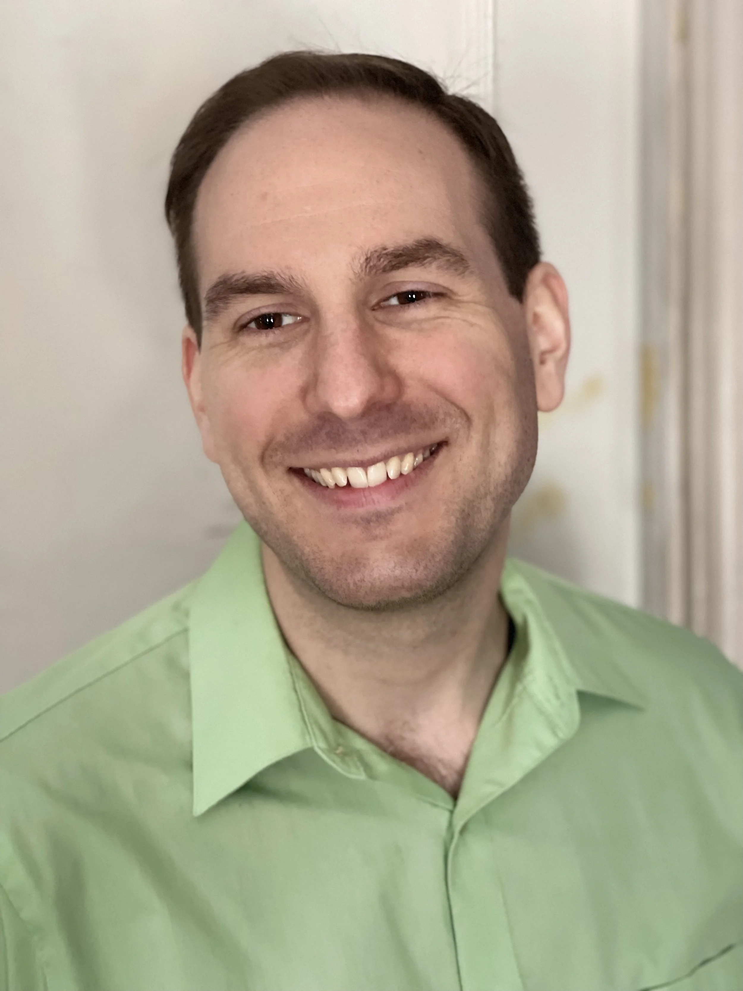A smiling man with dark hair and light skin wearing a light green collared shirt.