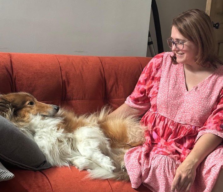 A woman in a pink patterned dress sitting on a red couch, smiling and interacting with a dog lying across her lap.