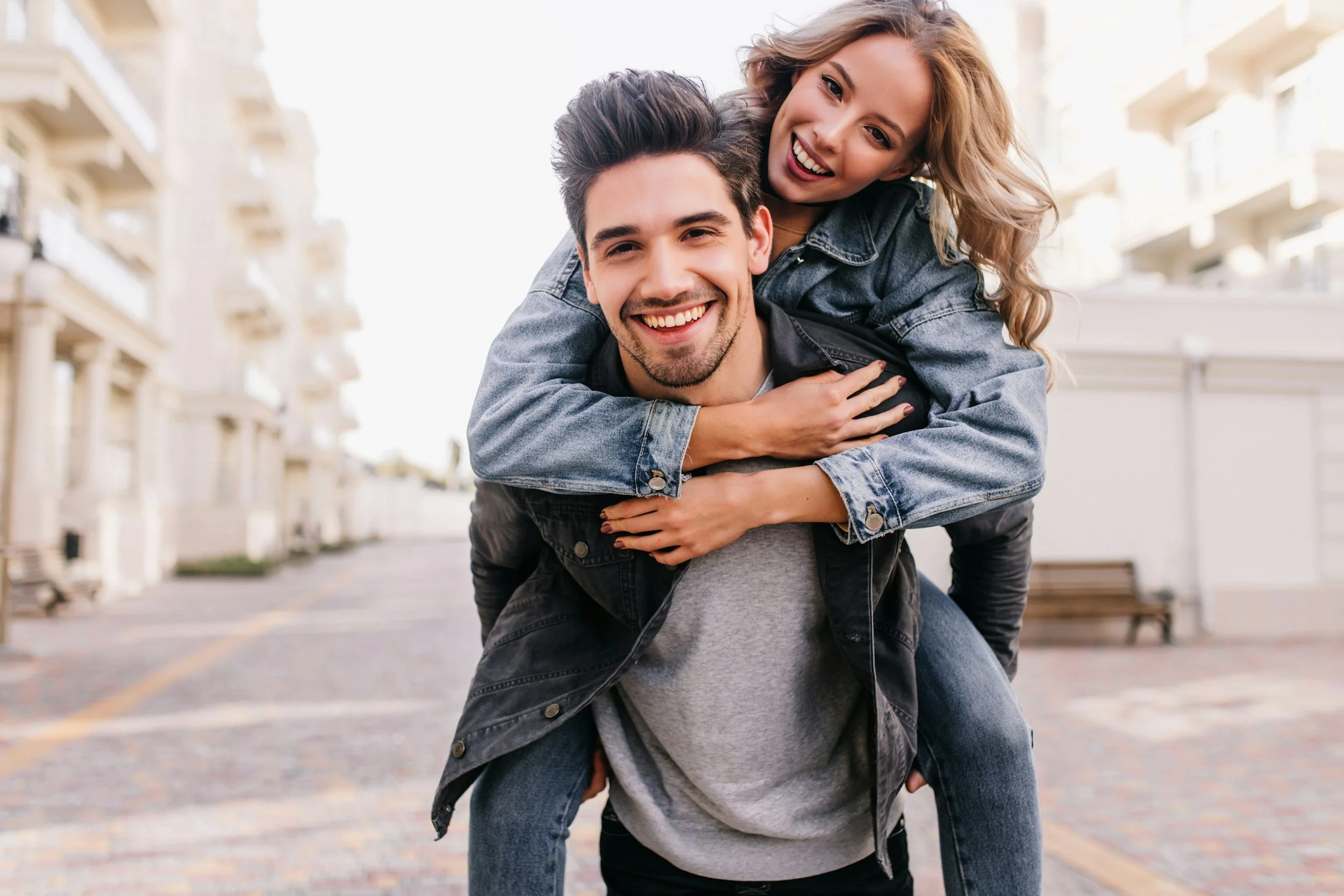 A happy young woman with blonde hair and a man with dark hair giving a piggyback ride in an outdoor urban setting with buildings in the background.