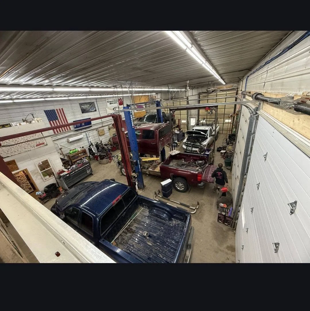 Interior of a large garage workshop with multiple vehicles, including trucks and cars, and various tools and equipment.