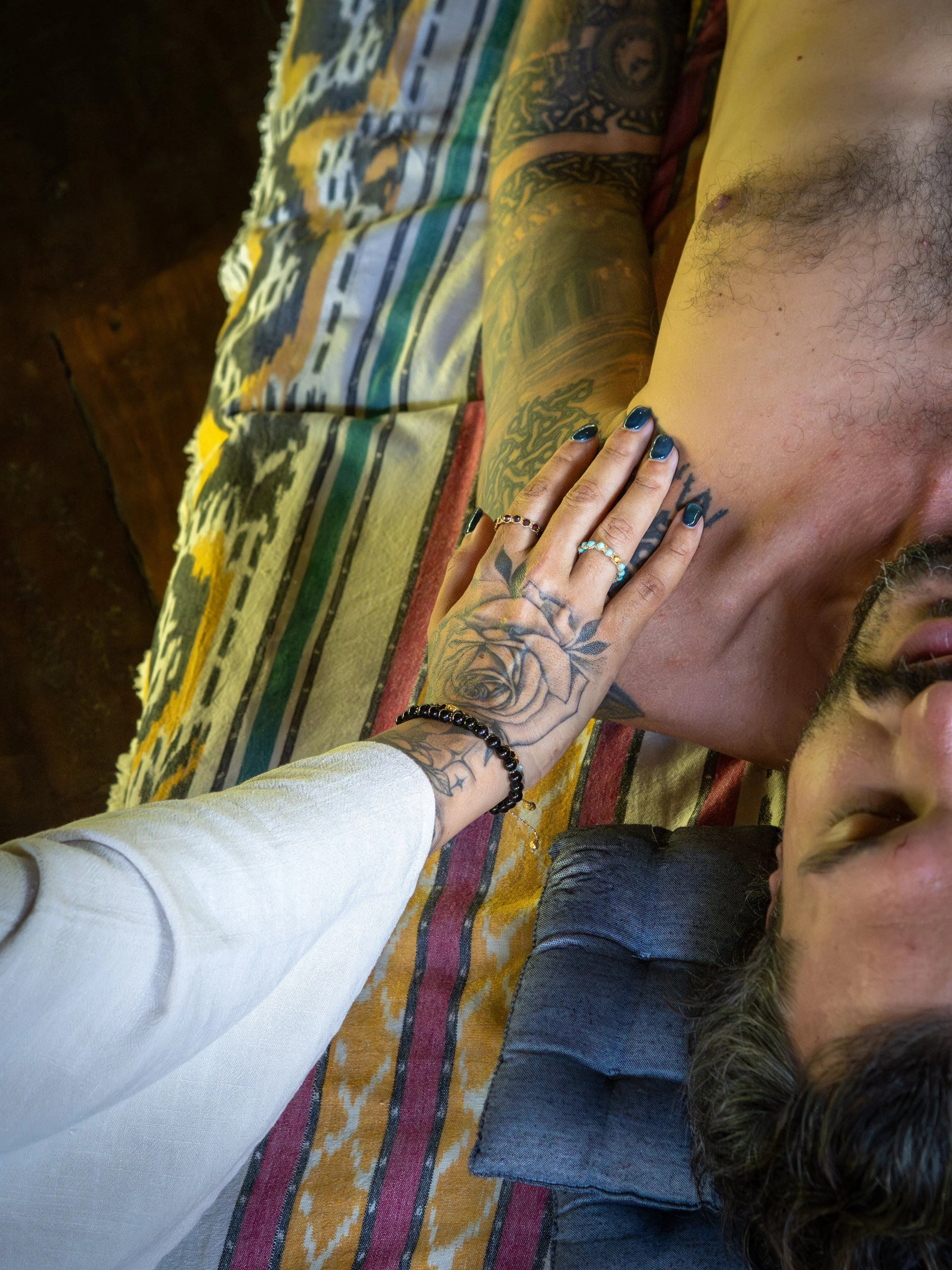 A tattooed man lying on a colorful woven blanket with a woman gently touching his chest. The man has dark curly hair, and the woman has long, dark, manicured nails and is wearing rings and a bracelet.