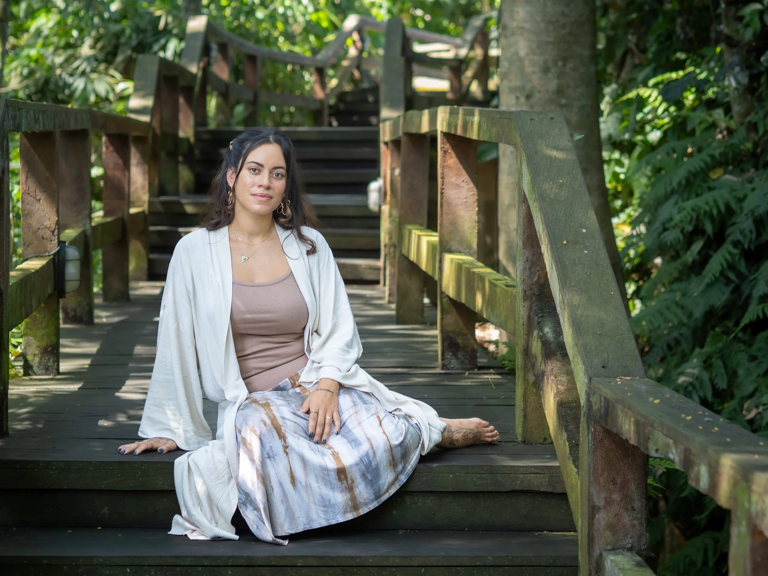 A woman sitting on a wooden pathway surrounded by green foliage, with stairs behind her, wearing a beige top, white cardigan, and patterned pants, with tattoos and jewelry.