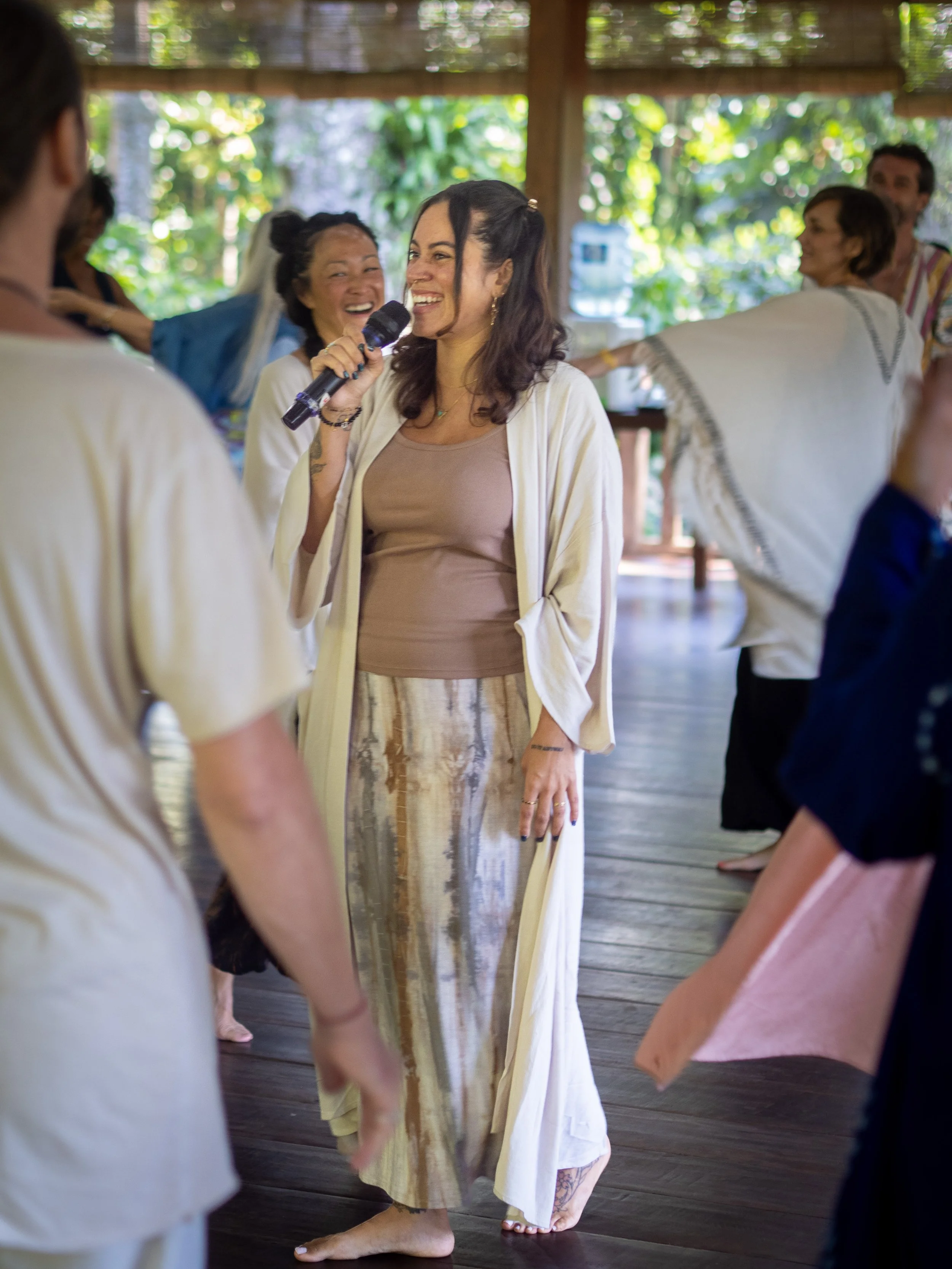A woman with dark hair and a tattoo on her ankle, holding a microphone, is smiling and participating in a dance or group activity indoors with other people. The setting has natural light and green foliage visible outside.