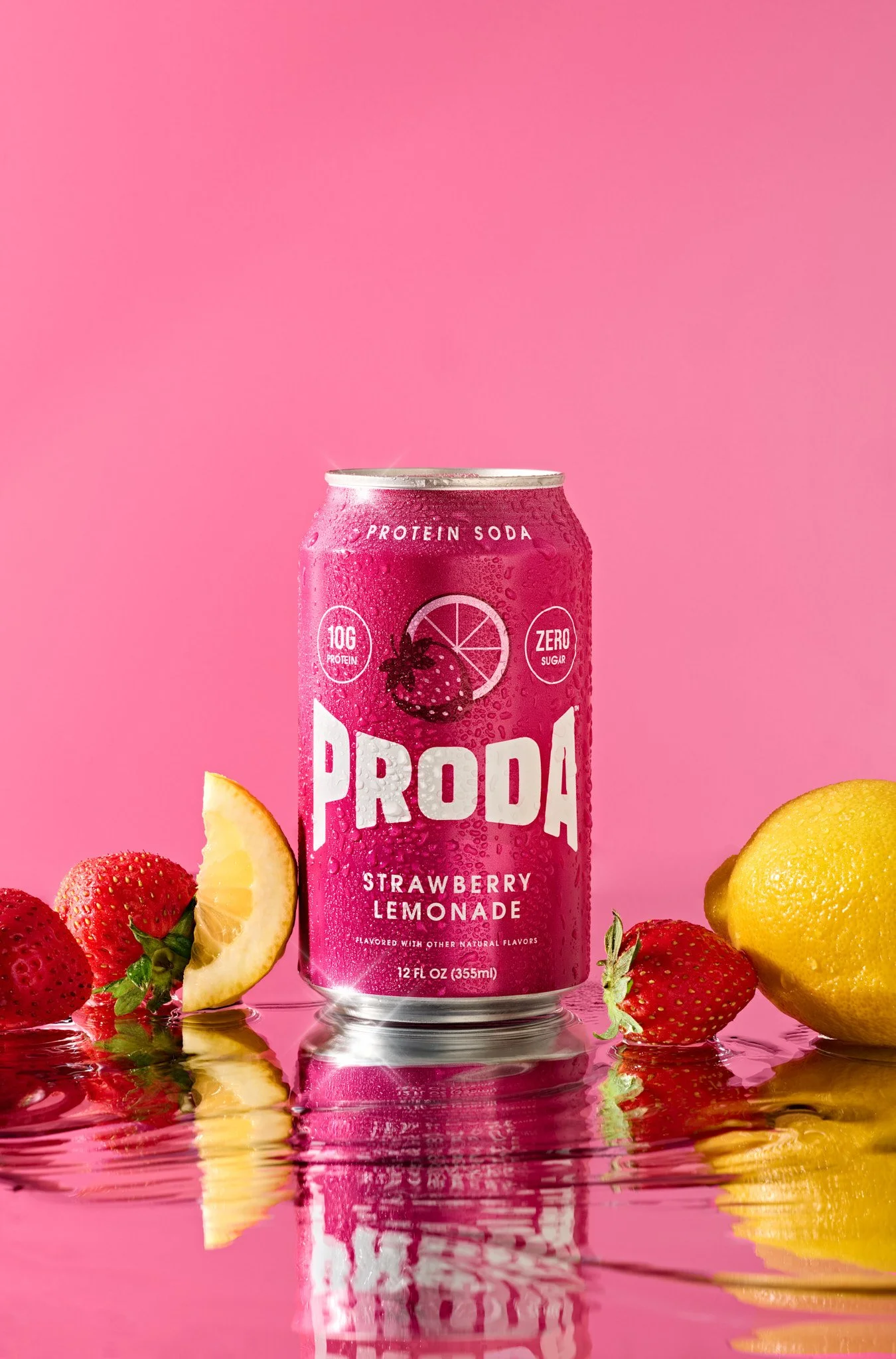 A pink can of protein soda with strawberries and lemon slices around it, reflecting on a water surface against a pink background. Pink protein soda can photographed with strawberry lemonade styling