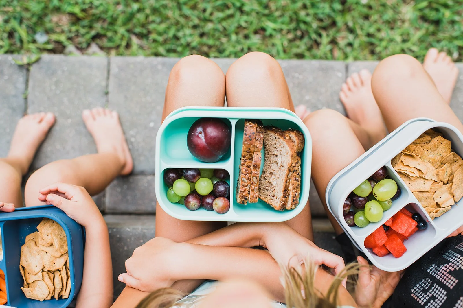 Children and adults sitting on a porch with lunch containers containing grapes, bread, watermelon, crackers, and an apple. small local business orlando product photography lifestyle