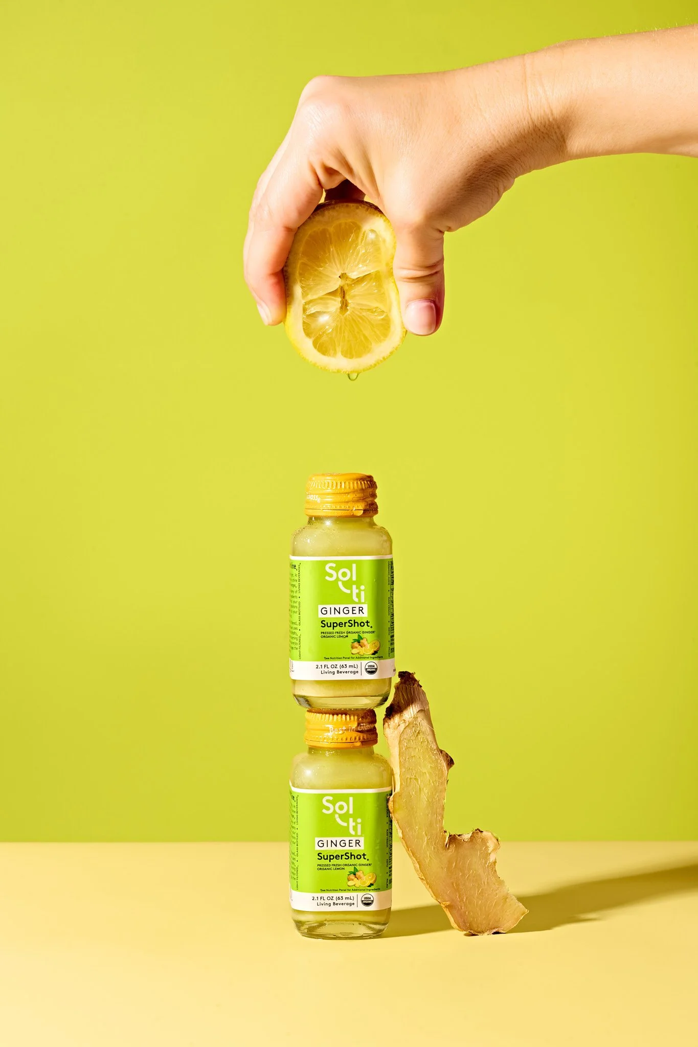 A clear bottle of Solti Ginger SuperShot placed on a yellow surface against a yellow background, with a hand squeezing a lemon half and a ginger piece in the scene. Colorful beverage product photography with condensation on glass