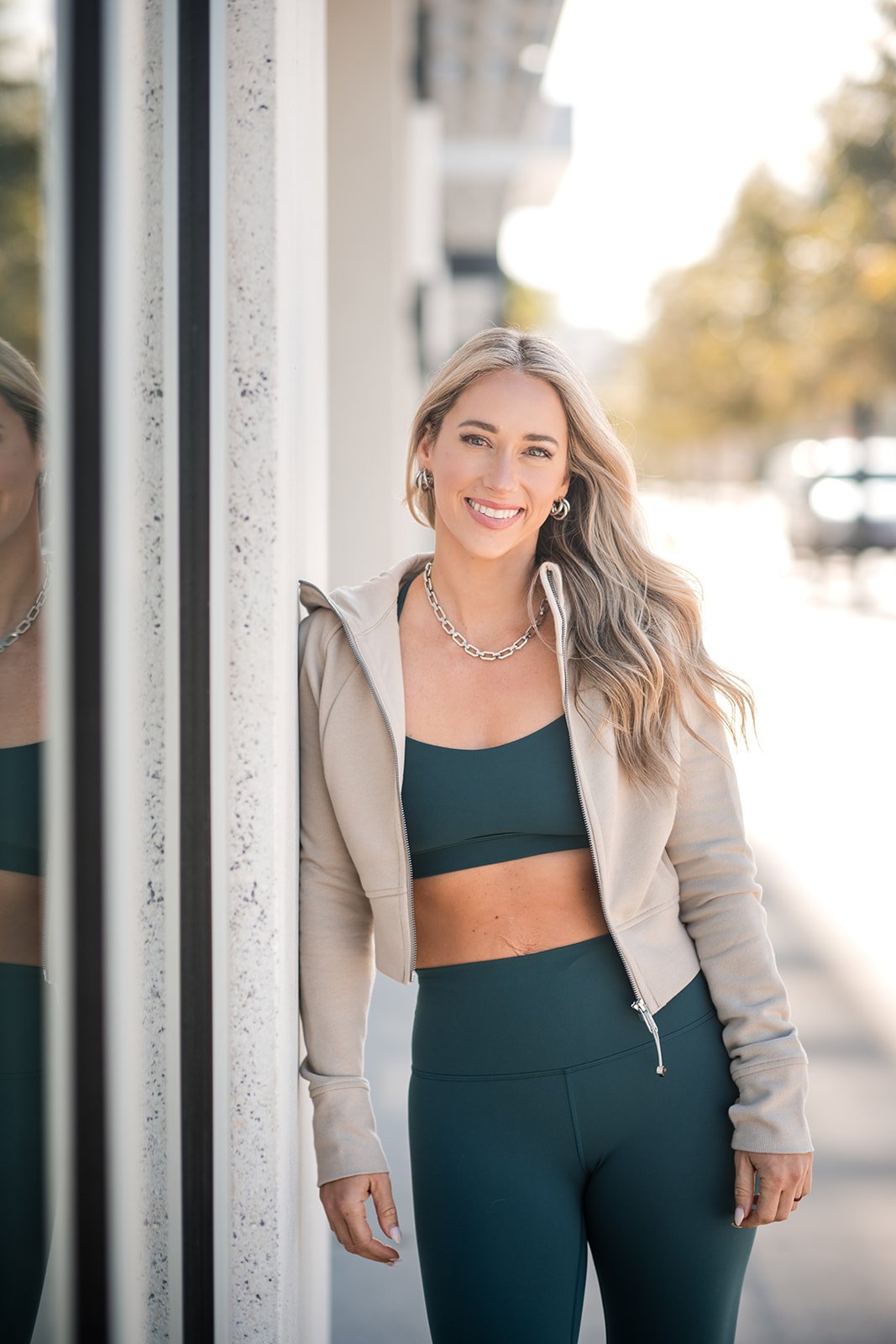 A woman with long wavy blonde hair smiling while standing outdoors next to a wall with a mirror, wearing a black sports bra, matching leggings, a beige jacket, silver jewelry, and natural makeup.