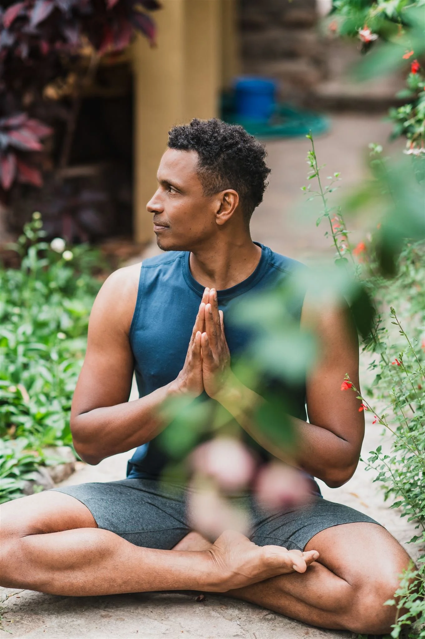 A man practicing yoga outdoors, sitting in a cross-legged position with hands in prayer position, surrounded by greenery.