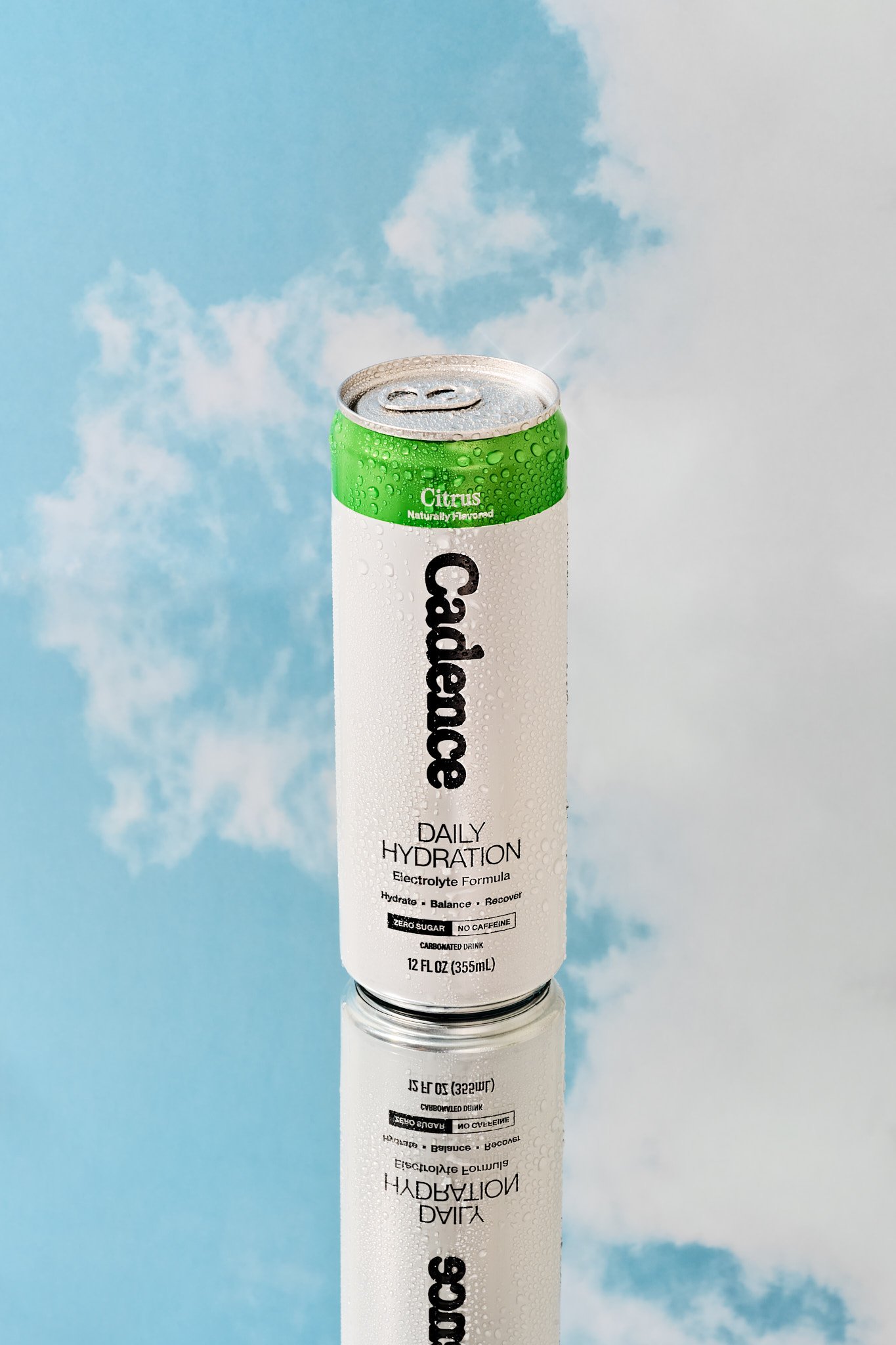 A can of citrus flavored electrolytes with droplets of condensation on its surface, positioned in front of a blue sky with scattered white clouds, with its reflection visible on a shiny surface below. Commercial beverage photography for a wellness an