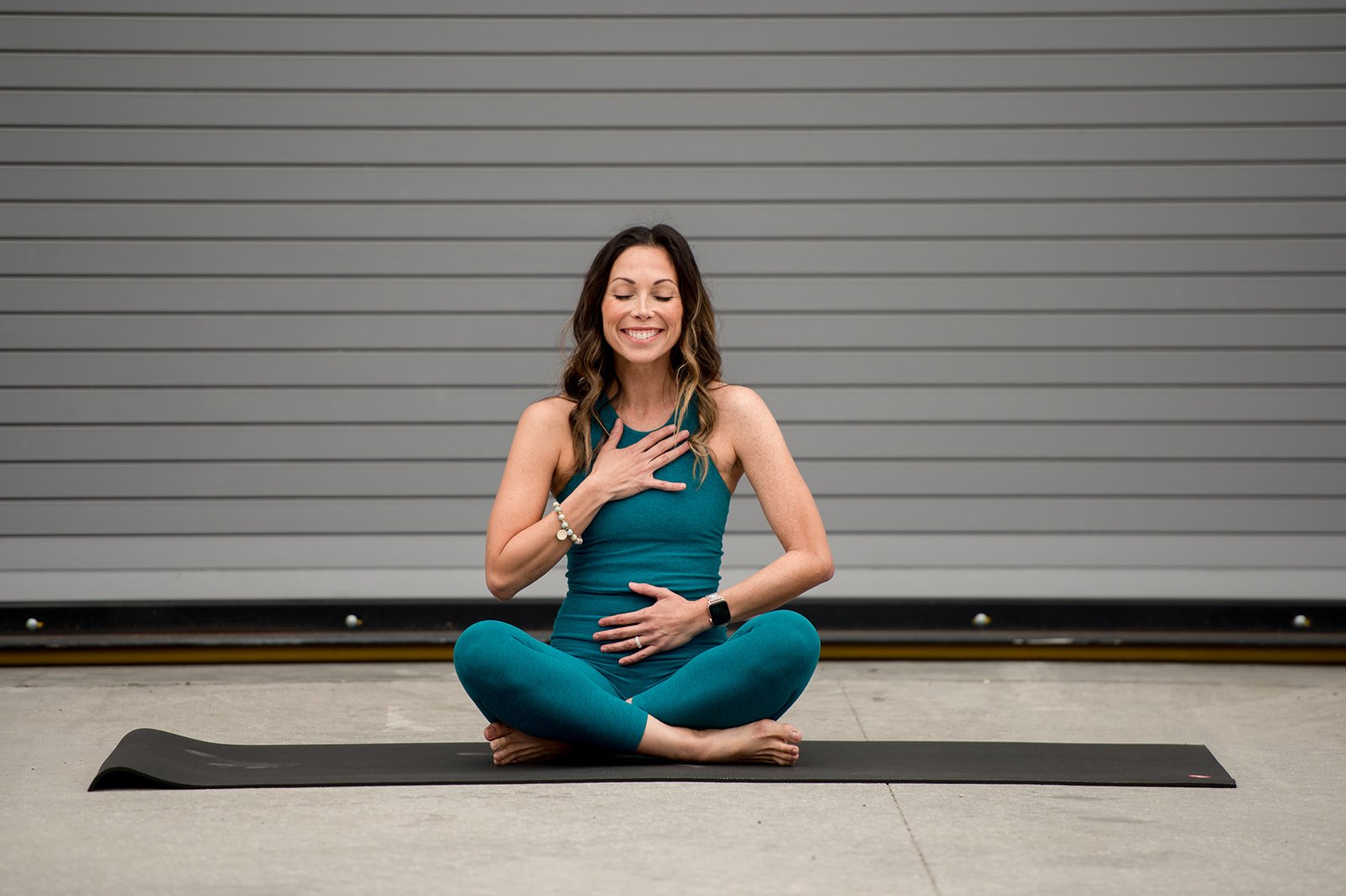 A woman practicing yoga in a seated meditation pose on a black mat outside, wearing teal workout clothes, with her eyes closed and a gentle smile, against a gray industrial background.