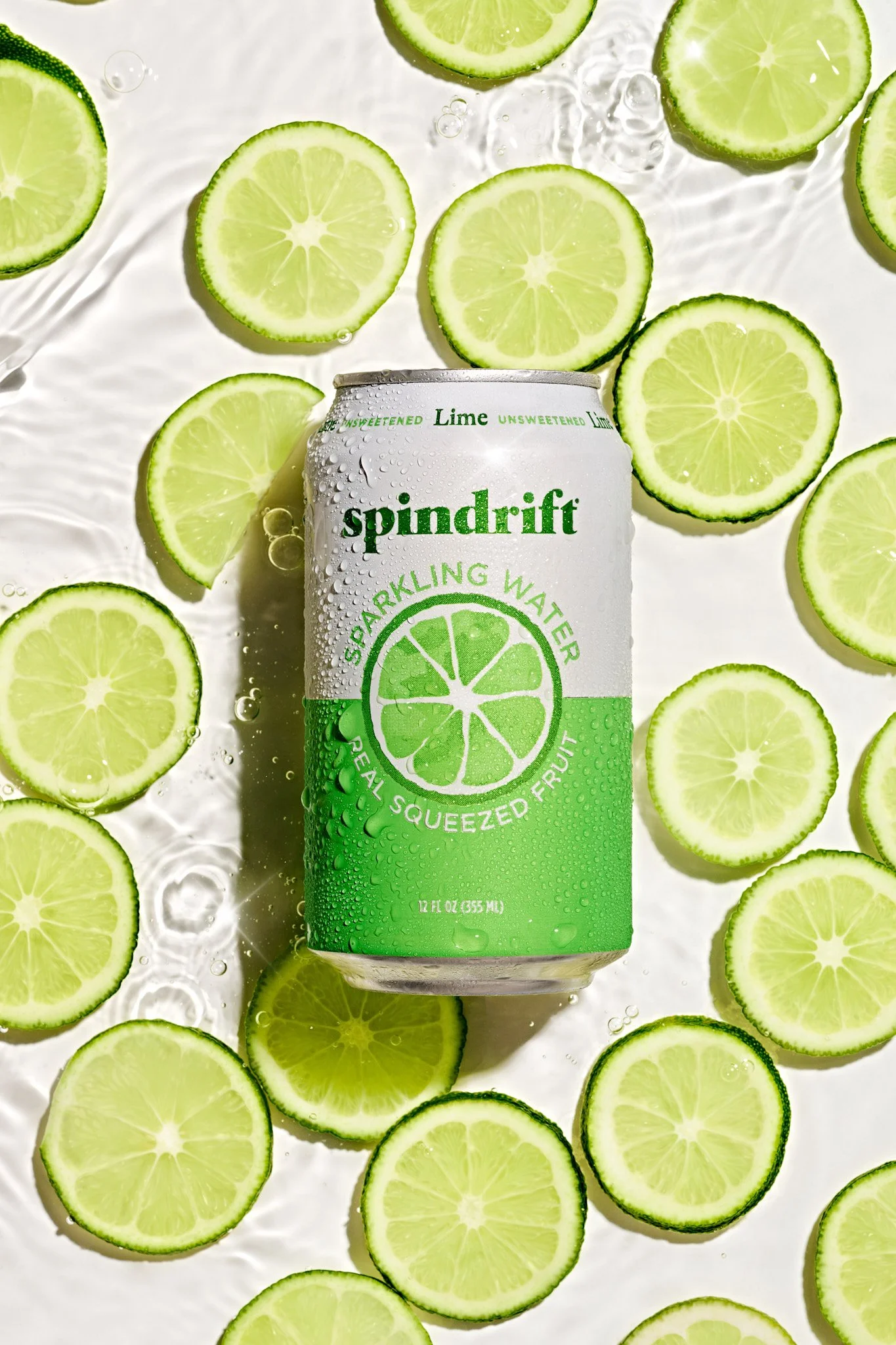 A can of SpinDrift sparkling water lemon flavor surrounded by sliced limes, with some slices partially submerged in water. Fitness lifestyle photography featuring a hydration product and runner
