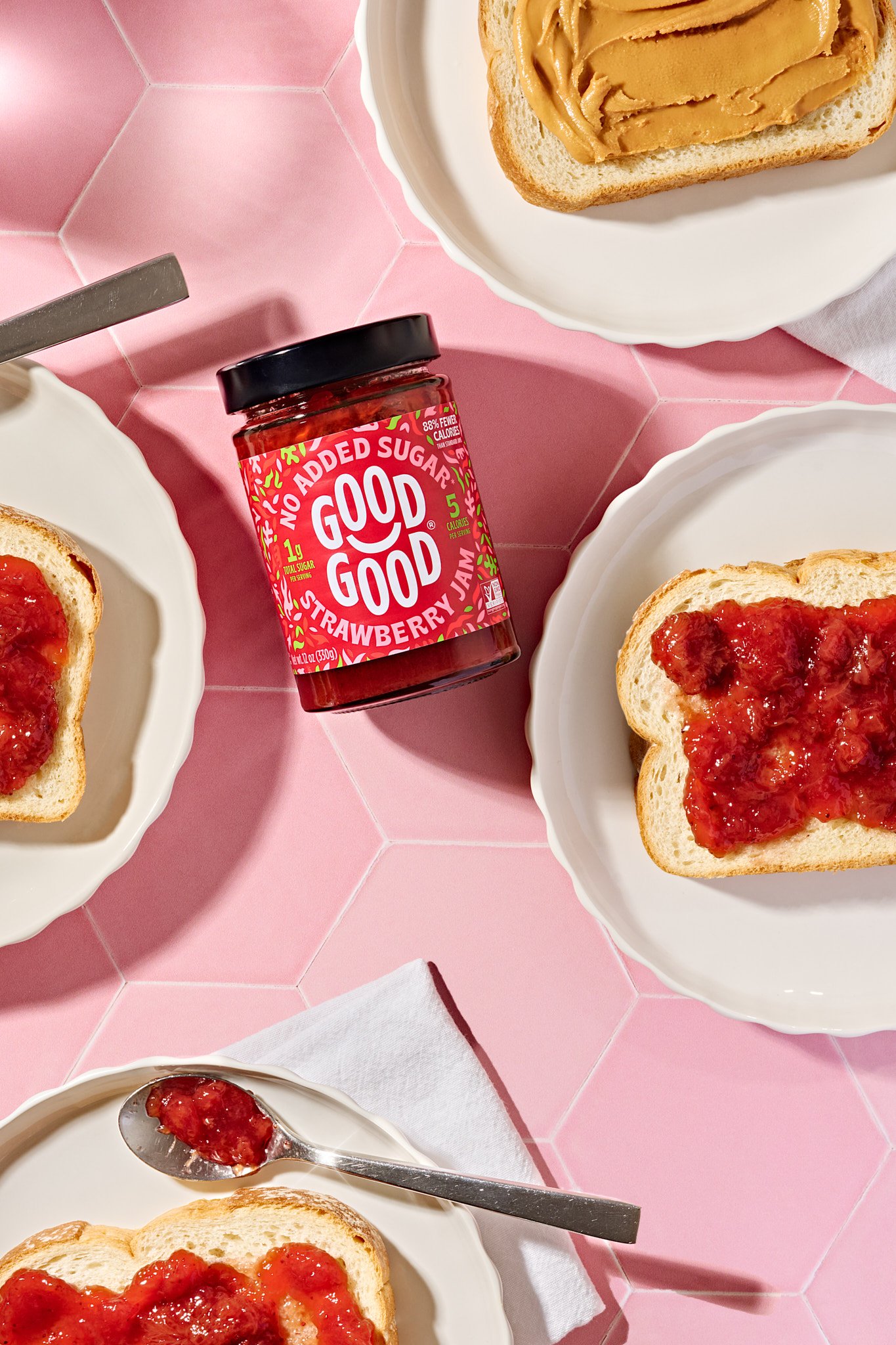 Slices of bread with strawberry jam and peanut butter on a pink tiled table, with a jar of strawberry jam nearby and a spoon with jam. Packaged food product photography for a modern consumer brand