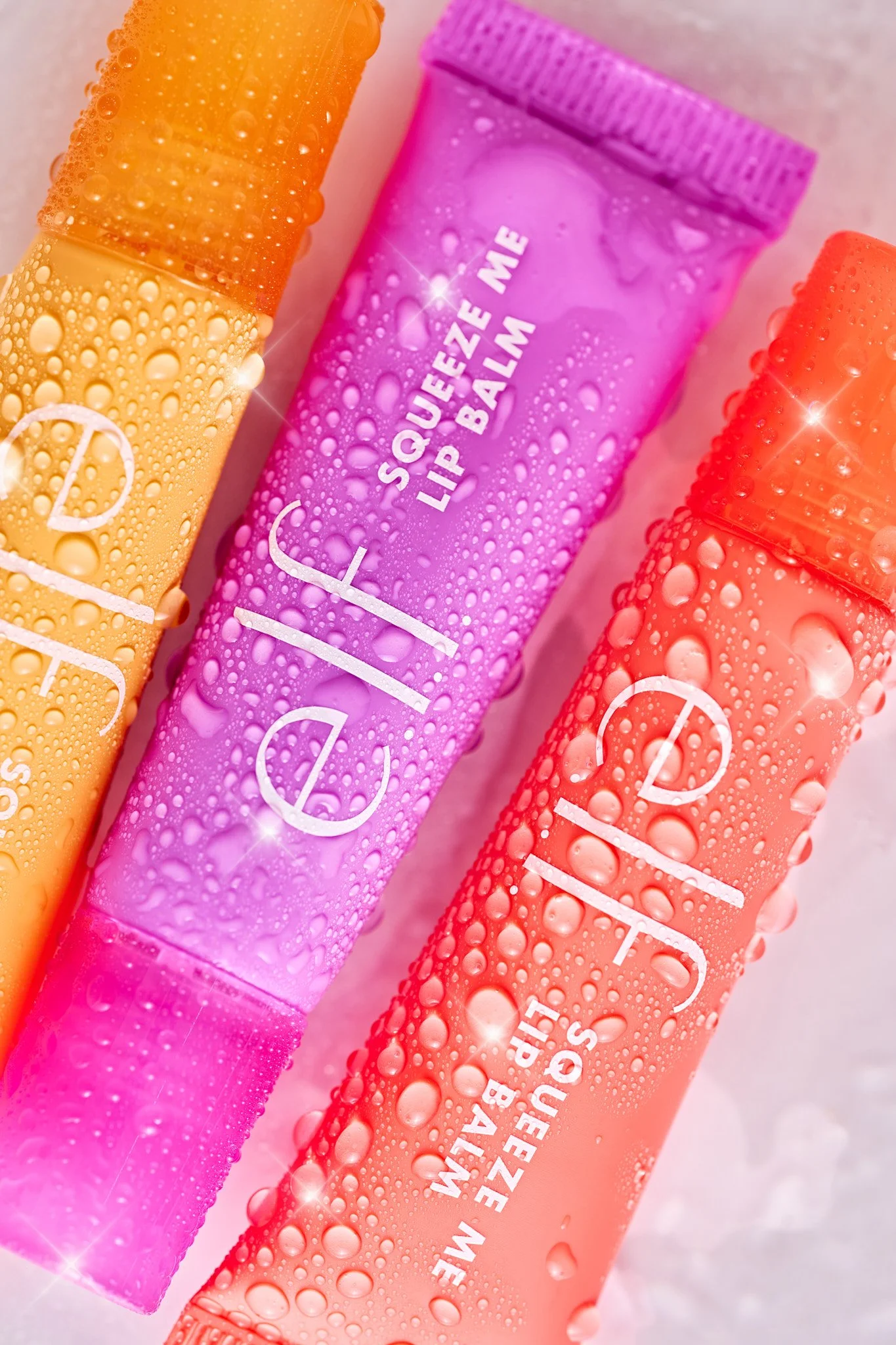 Close-up of three colorful lip balm tubes with water droplets on them, in orange, pink, and peach colors, positioned on a light background. For elf cosmetics beauty brand creative studio photography for CPG brands.