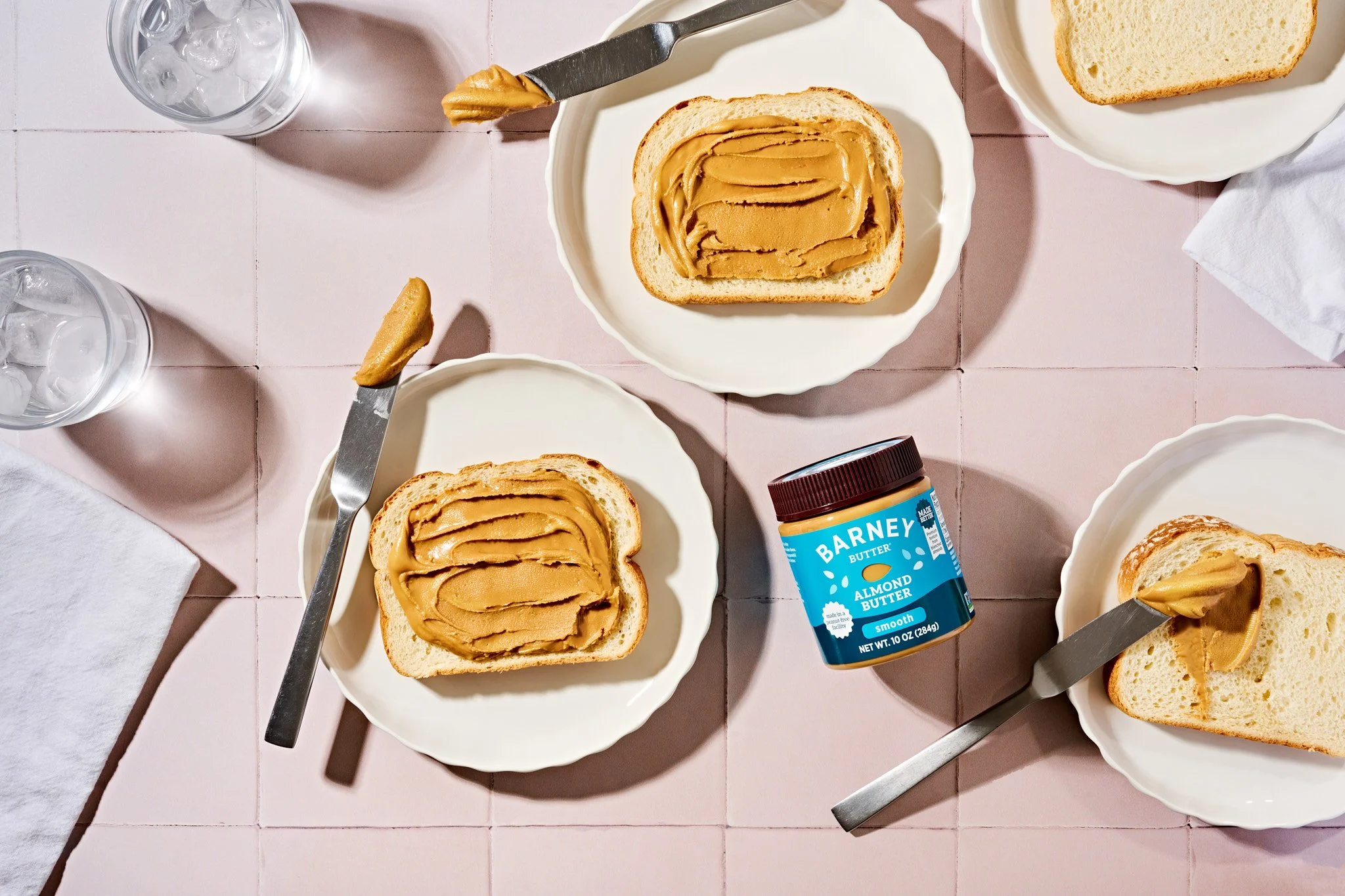 Three slices of bread with peanut butter on white plates, two glasses of ice water, and a jar of Barney Almond Butter on a pink tiled table. Commercial food photographer creating colorful imagery