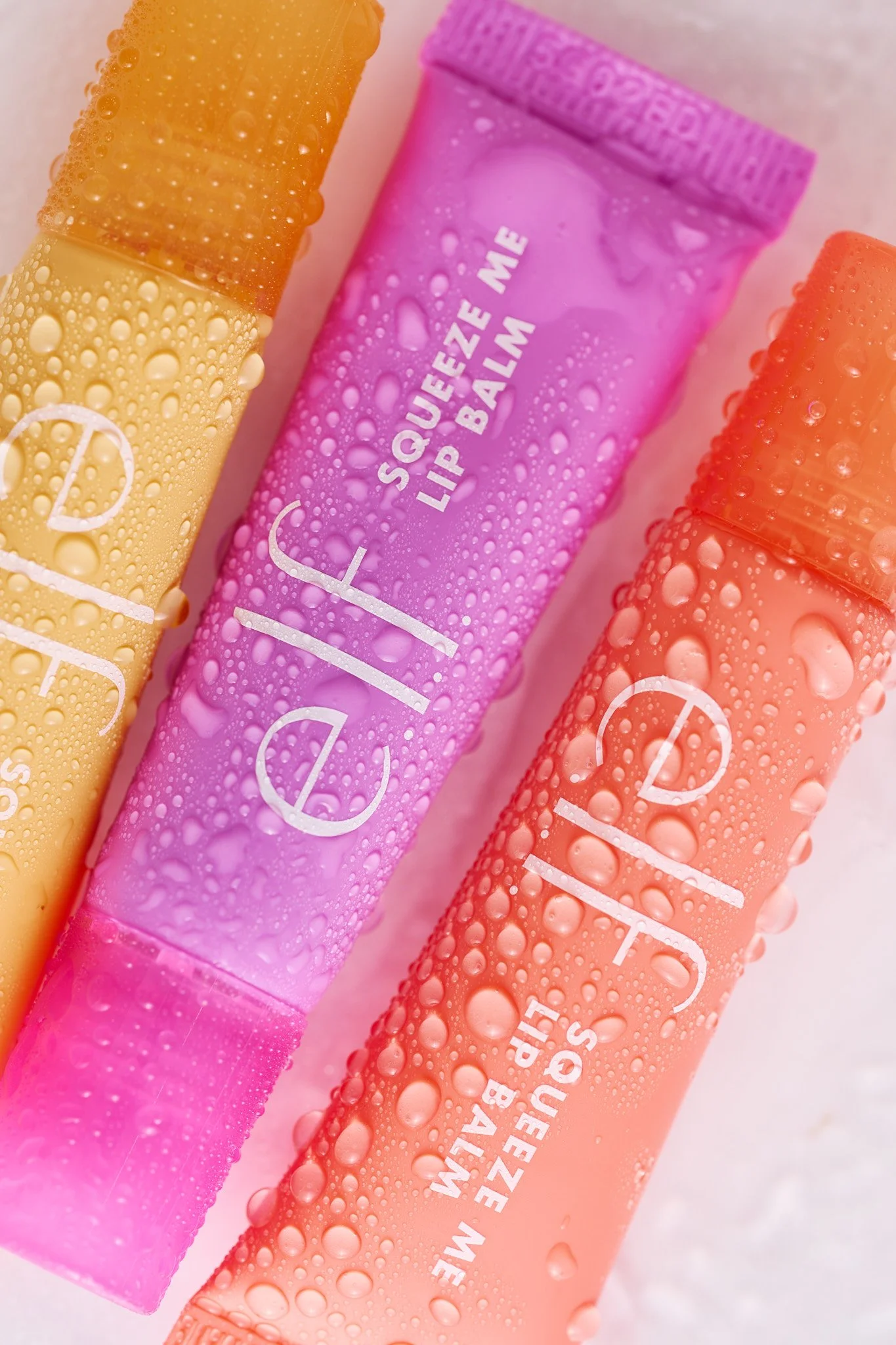 Close-up of three lip balm tubes in yellow, purple, and orange, covered in water droplets, with partially visible white text on each tube. vibrant beauty brand cpg photography