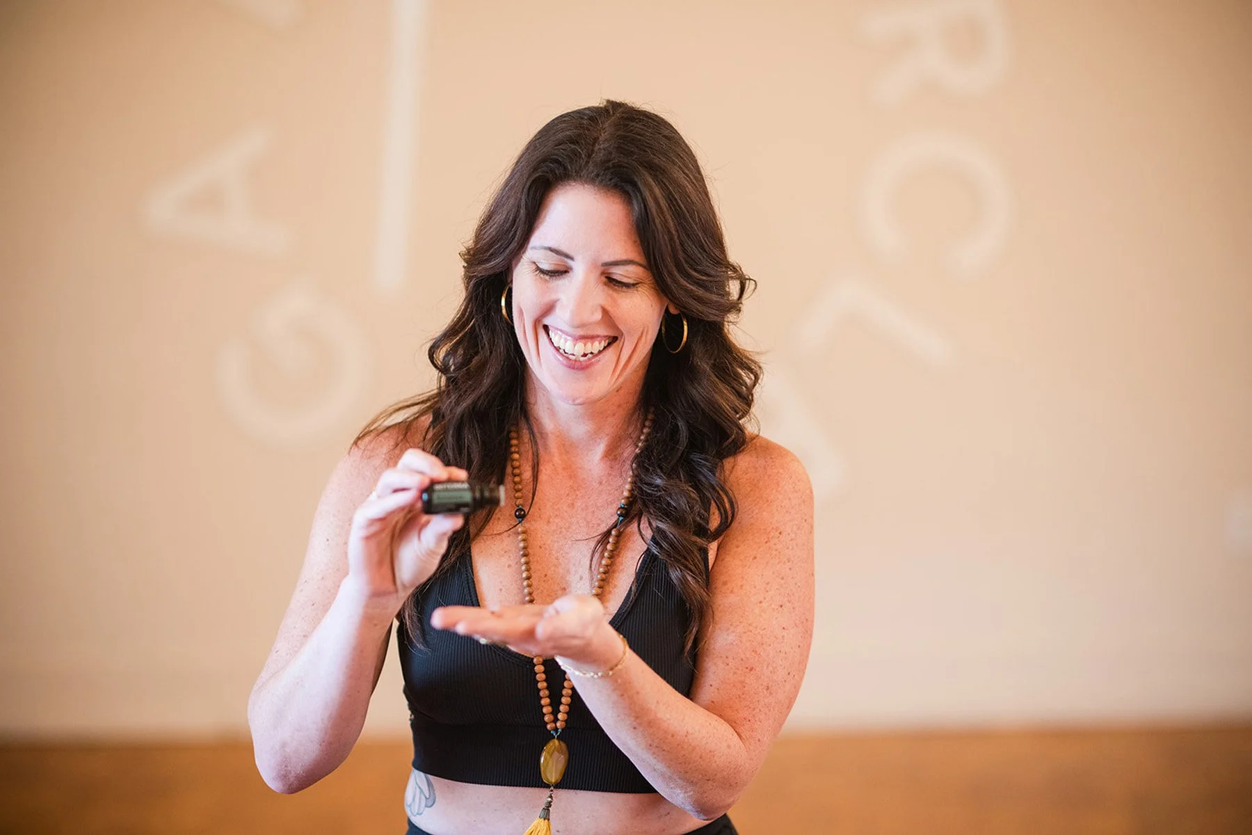 Woman with long, wavy brown hair smiling and looking down, holding and examining a small black object in her hand. yoga wellness essential oils doterra yoga studio