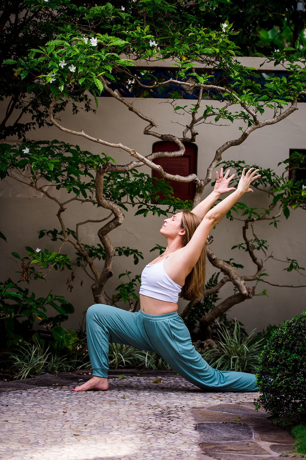A woman practicing yoga outdoors near a tree with white flowers, wearing a white tank top and teal pants, in a low lunge pose with arms raised.